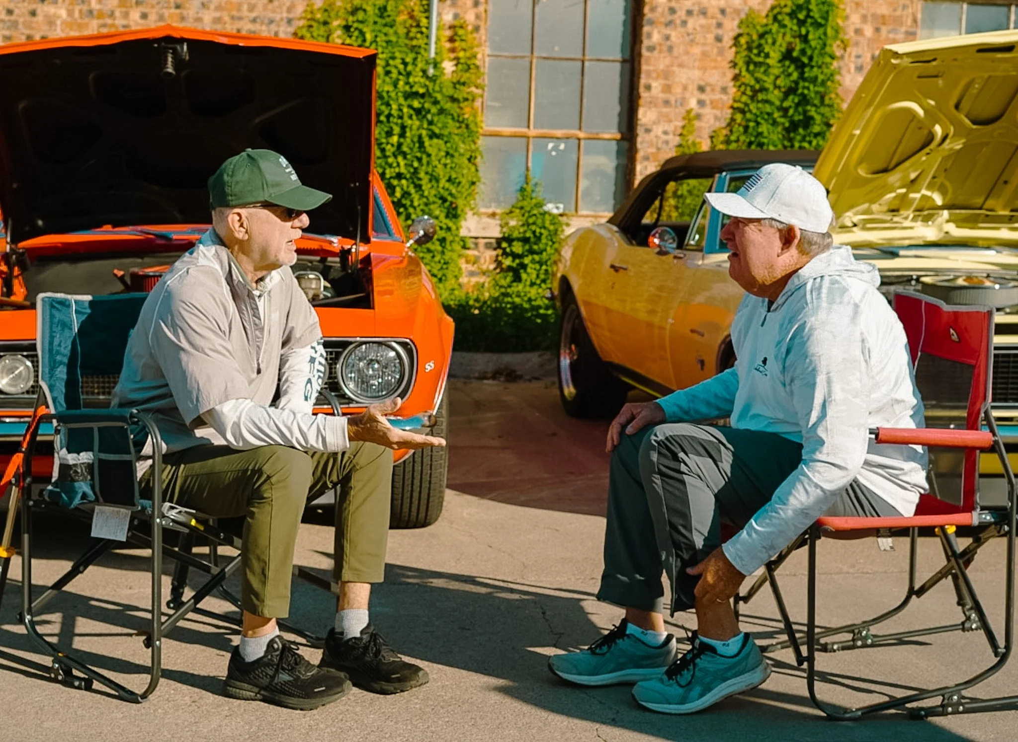 Two older men wearing caps are sitting and kneeling in front of classic cars with their hoods open, engaging in conversation outdoors near a brick building with green plants.