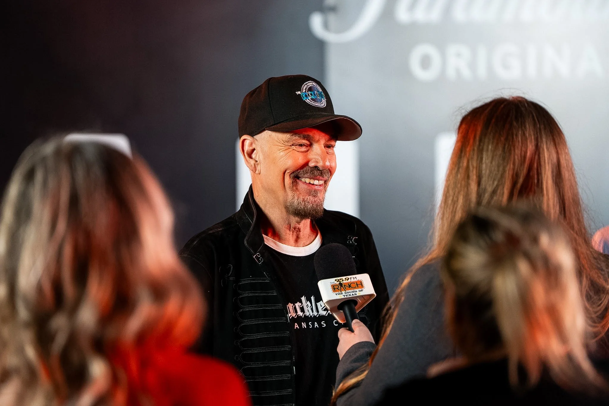Man smiling wearing a black cap speaking into a microphone at an event with a blurred audience in the foreground.