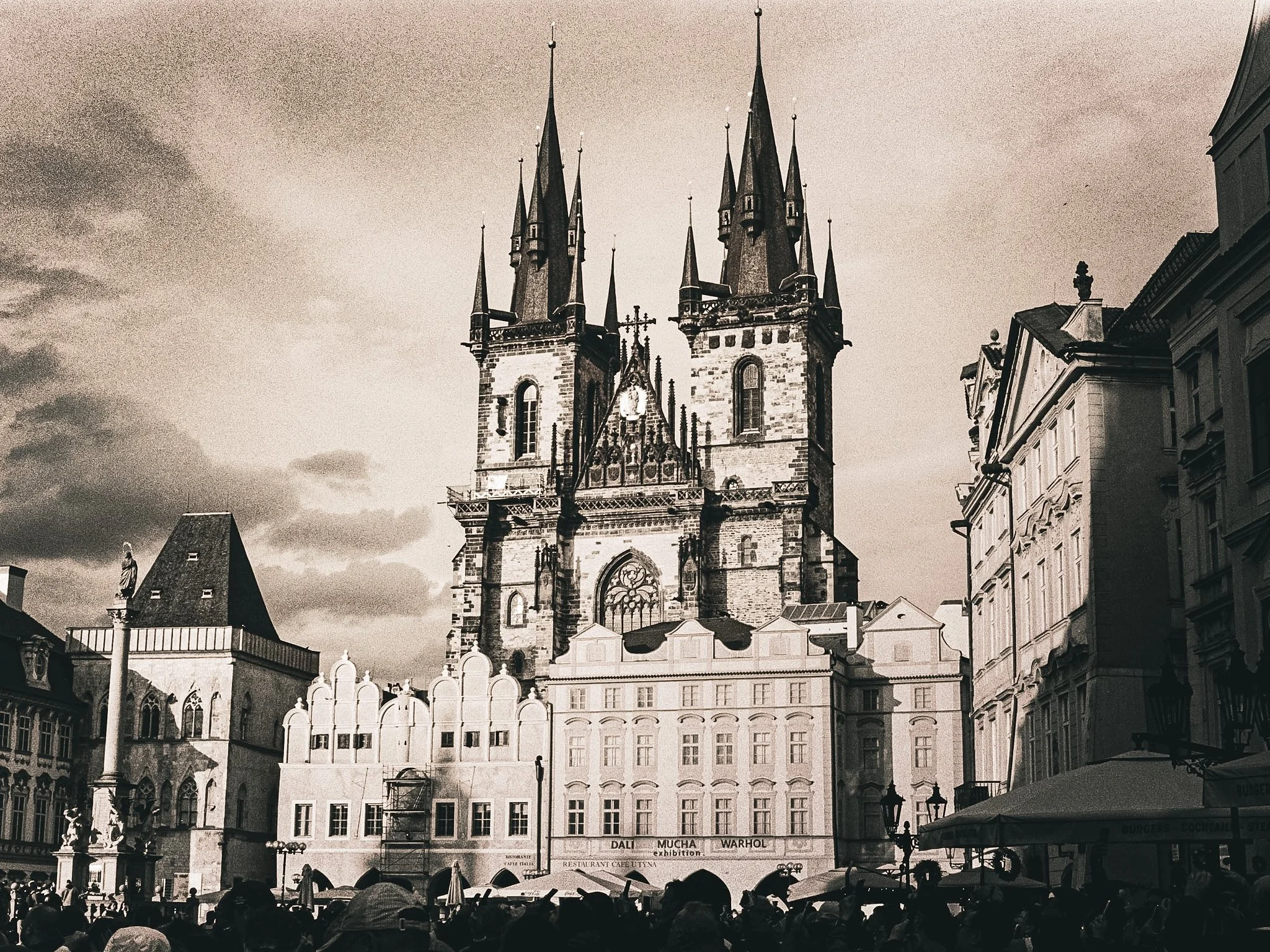 Black and white photo of a Gothic-style church with tall spires, surrounded by buildings and an outdoor cafe with umbrellas and people.