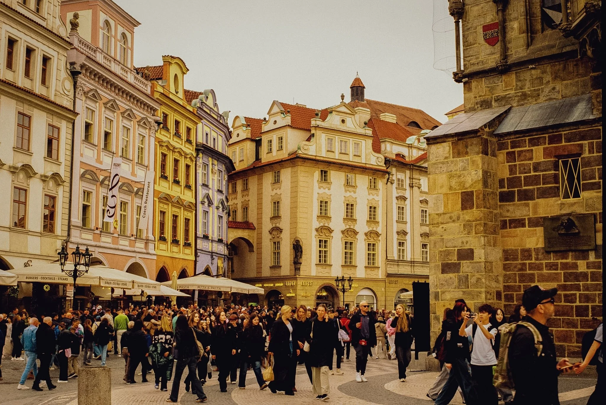 Crowded city square with historic buildings featuring colorful facades and red-tiled roofs, outdoor cafes with umbrellas, and many pedestrians walking or standing in the area.