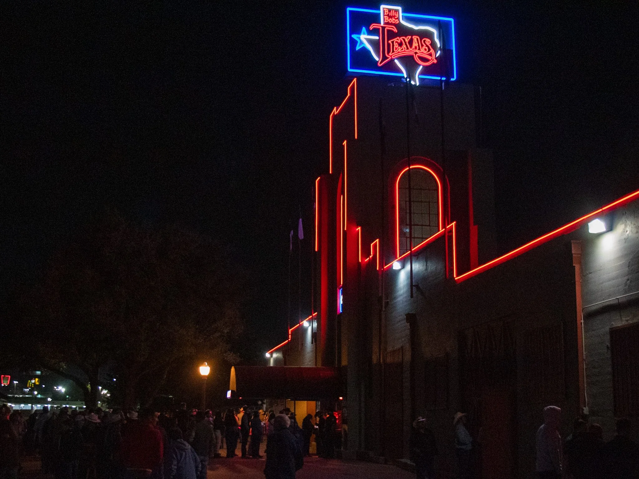Nighttime scene outside Billy Bob's Texas showing a crowd of people gathered near the building. The building features neon signs with red, blue, and white lights, including a large sign with the words 'Billy Bob's Texas' and an outline of the state o