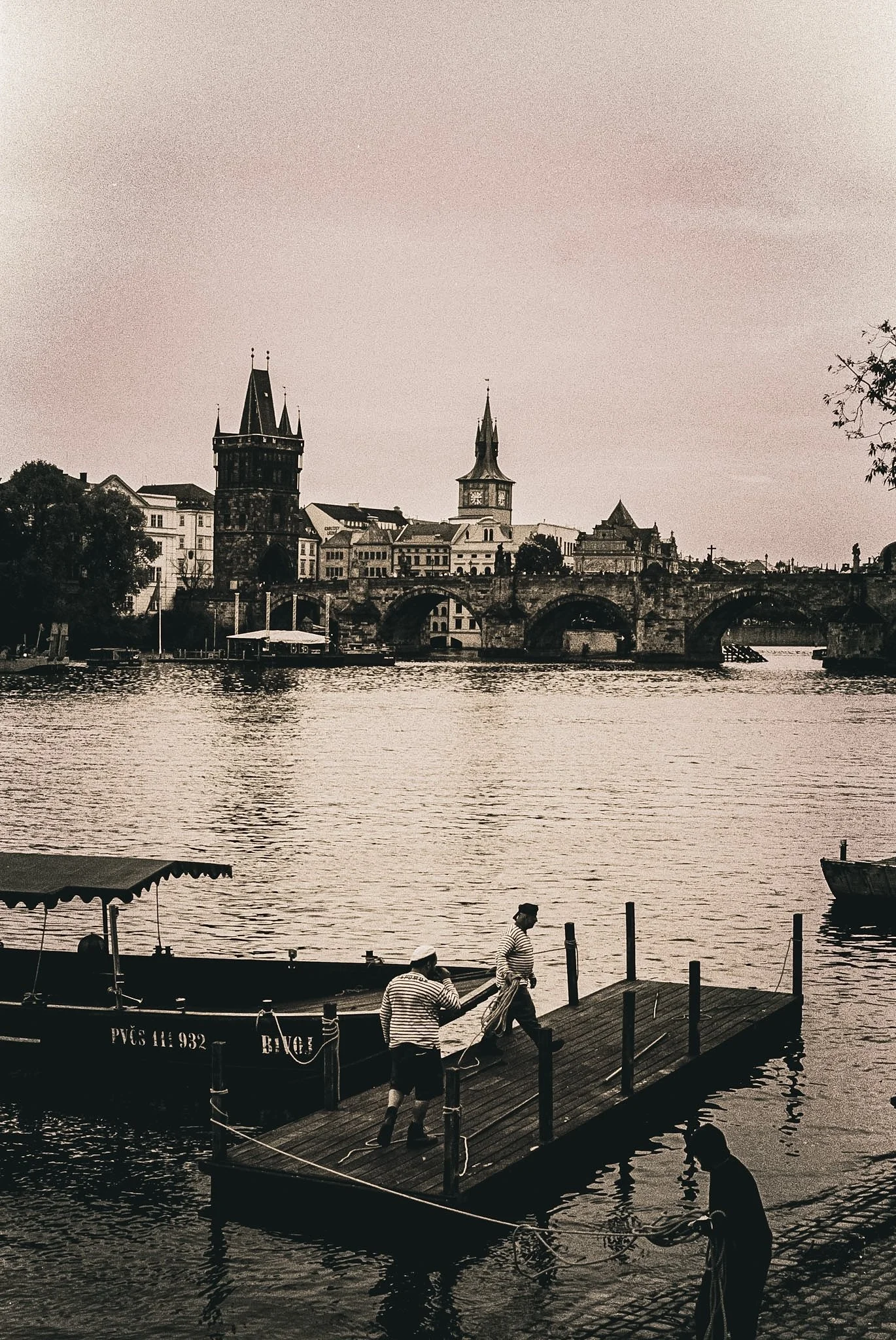 People working on yachts at a dock along the Charles River with historic buildings and bridges in the background in Prague, Czech Republic.