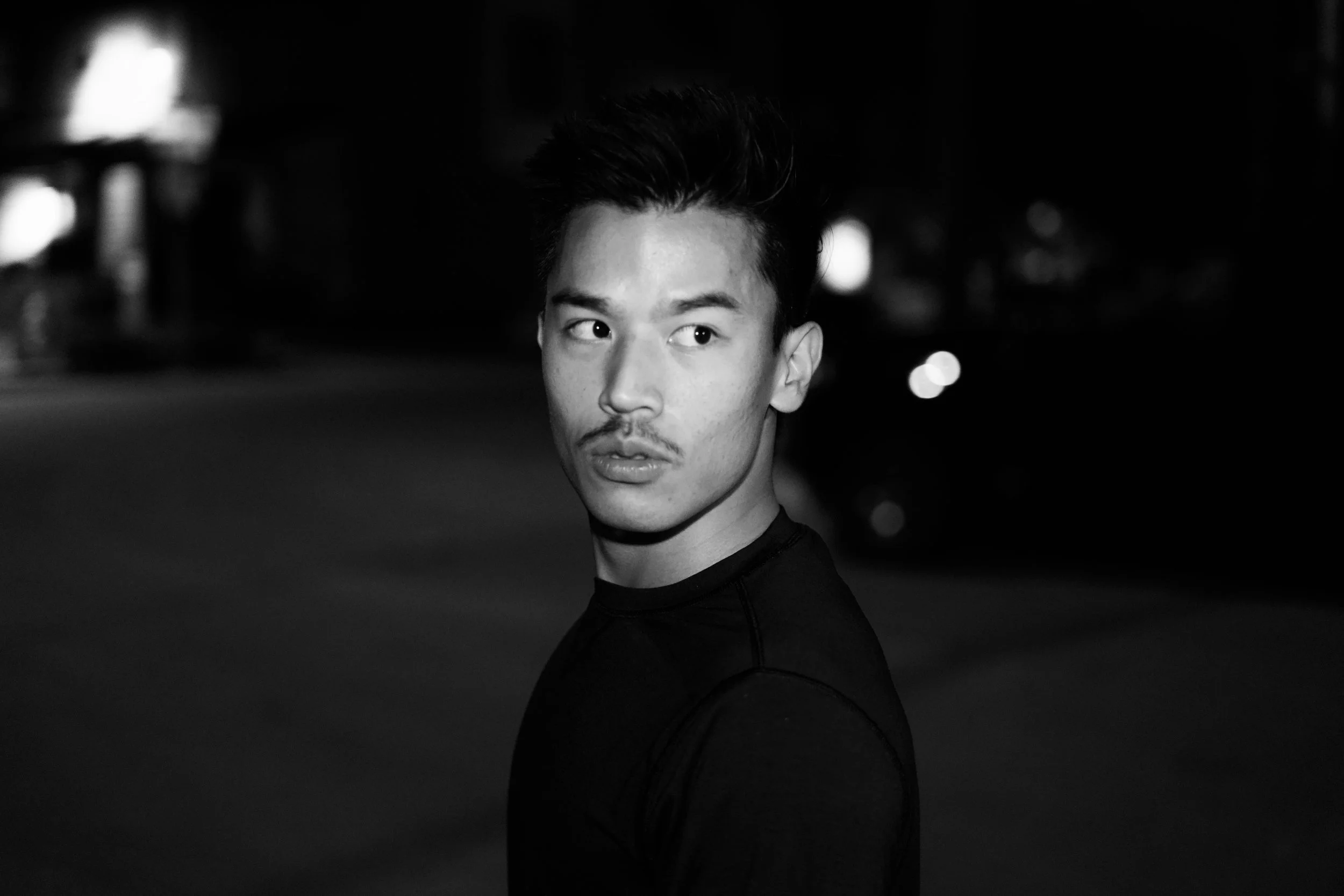 A black and white portrait of a young man with styled hair, looking over his shoulder at night in an urban setting with blurred lights in the background.