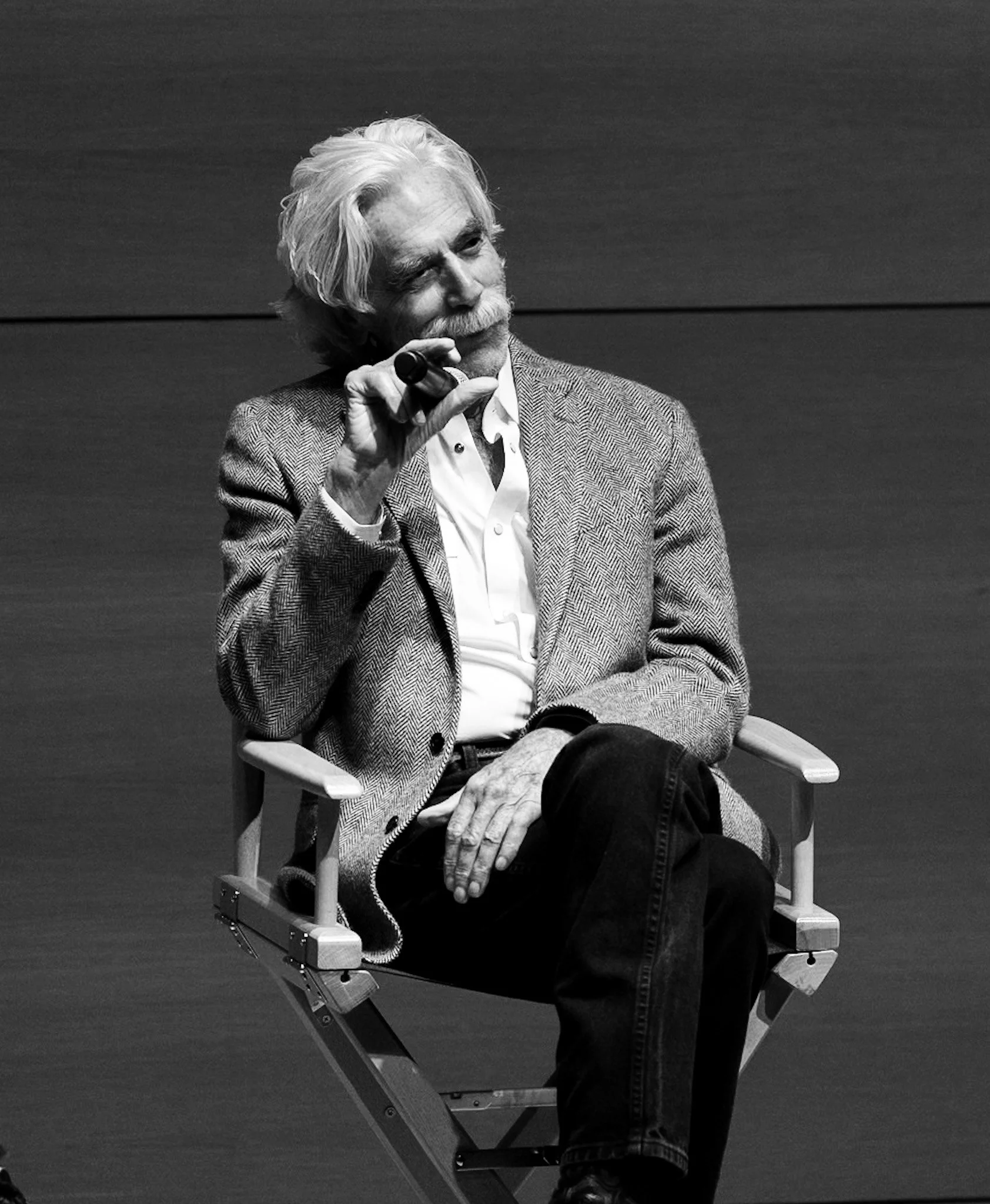 An elderly man with white hair and a beard, wearing a checkered blazer and white shirt, sitting on a chair with one leg crossed over the other. He has glasses hanging from his shirt and is holding a small device near his face. The background is plain