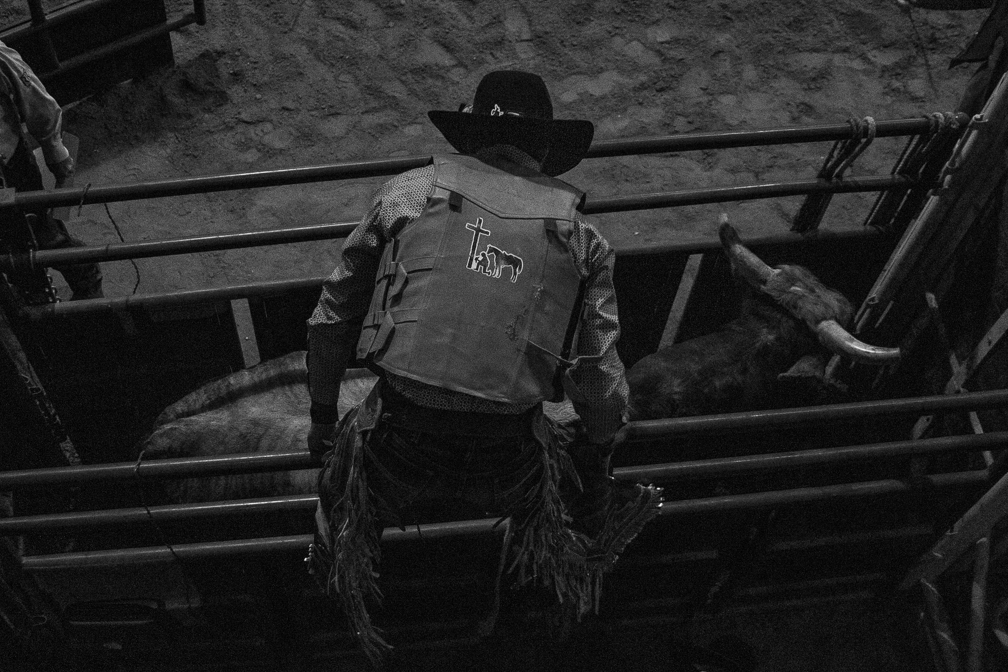 A person with a cowboy hat and a vest featuring a church and a bear logo, standing by a fence at a rodeo or horse riding arena, with dirt ground visible.