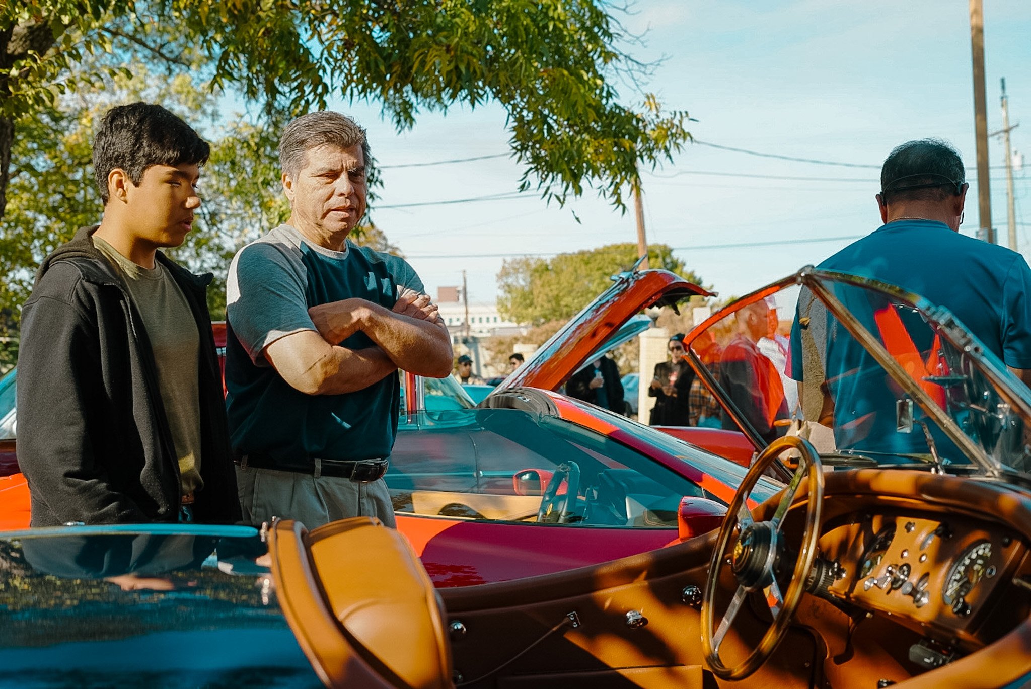 Three men observing a red vintage car at an outdoor car show with trees and power lines visible in the background.