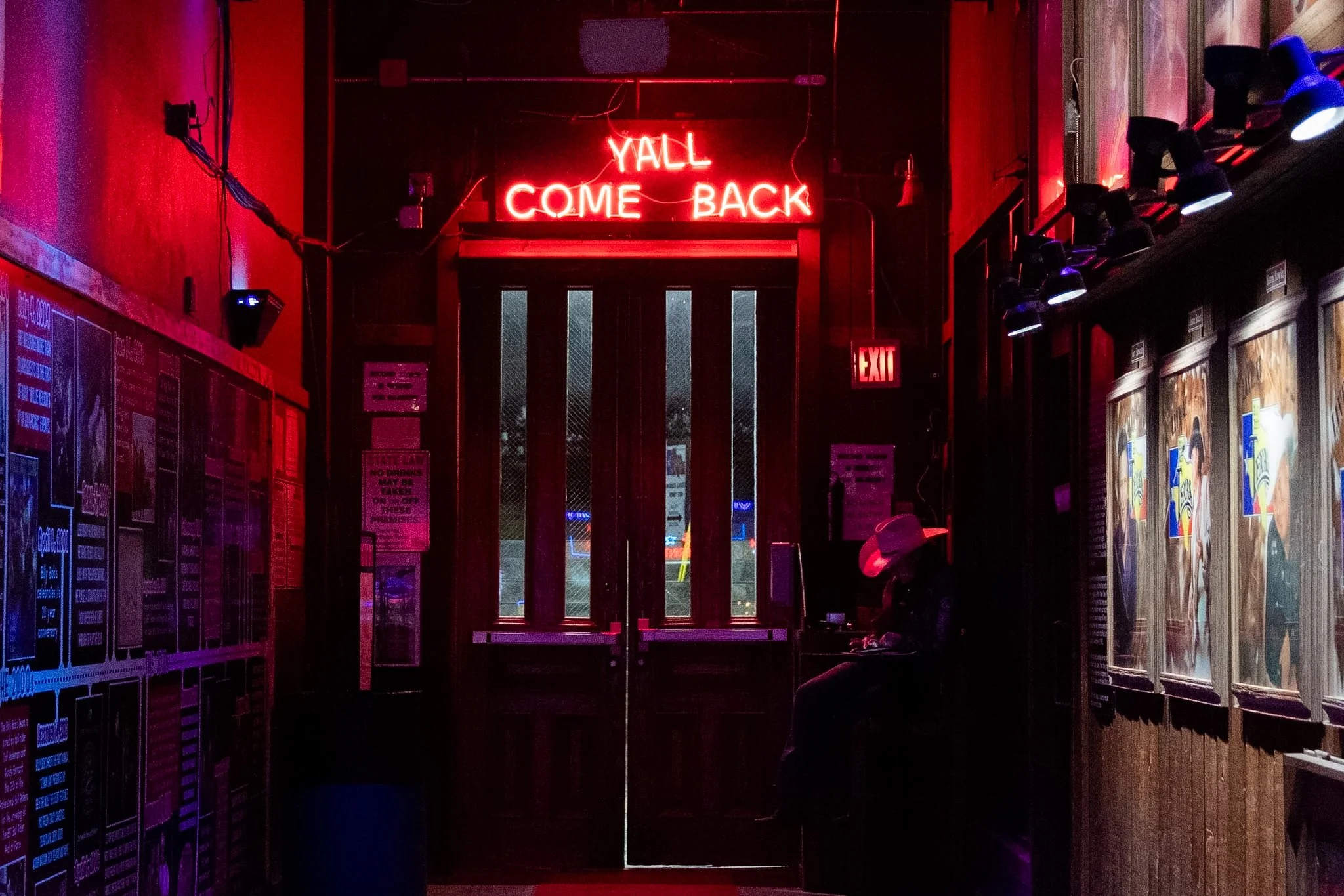 Neon sign reading 'YALL COME BACK' hanging above a door in a dimly lit bar or restaurant interior with red and purple lighting.