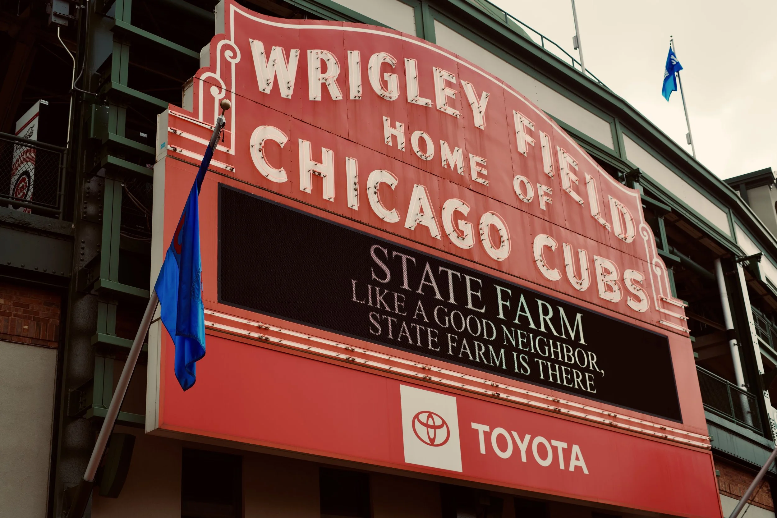 The image shows a large red billboard with white text that says 'WRIGLEY HOME OF CHICAGO CUBS'. Below, there is a digital sign with a message about the state farm and Toyota logos, and a flag is flying nearby. The background includes a green-toned bu