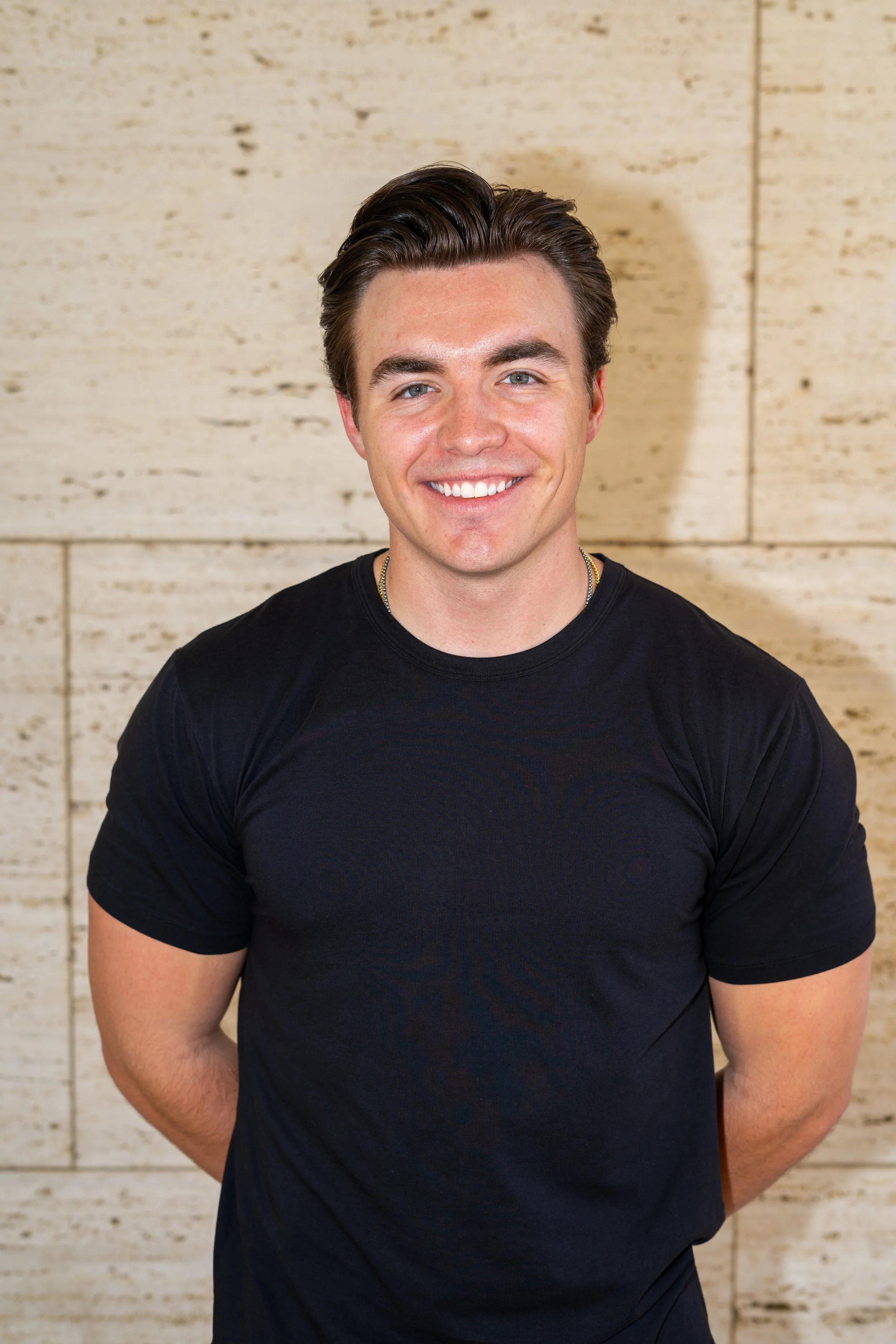 A young man with dark brown hair, blue eyes, and a fair complexion smiling in front of a light beige textured wall.