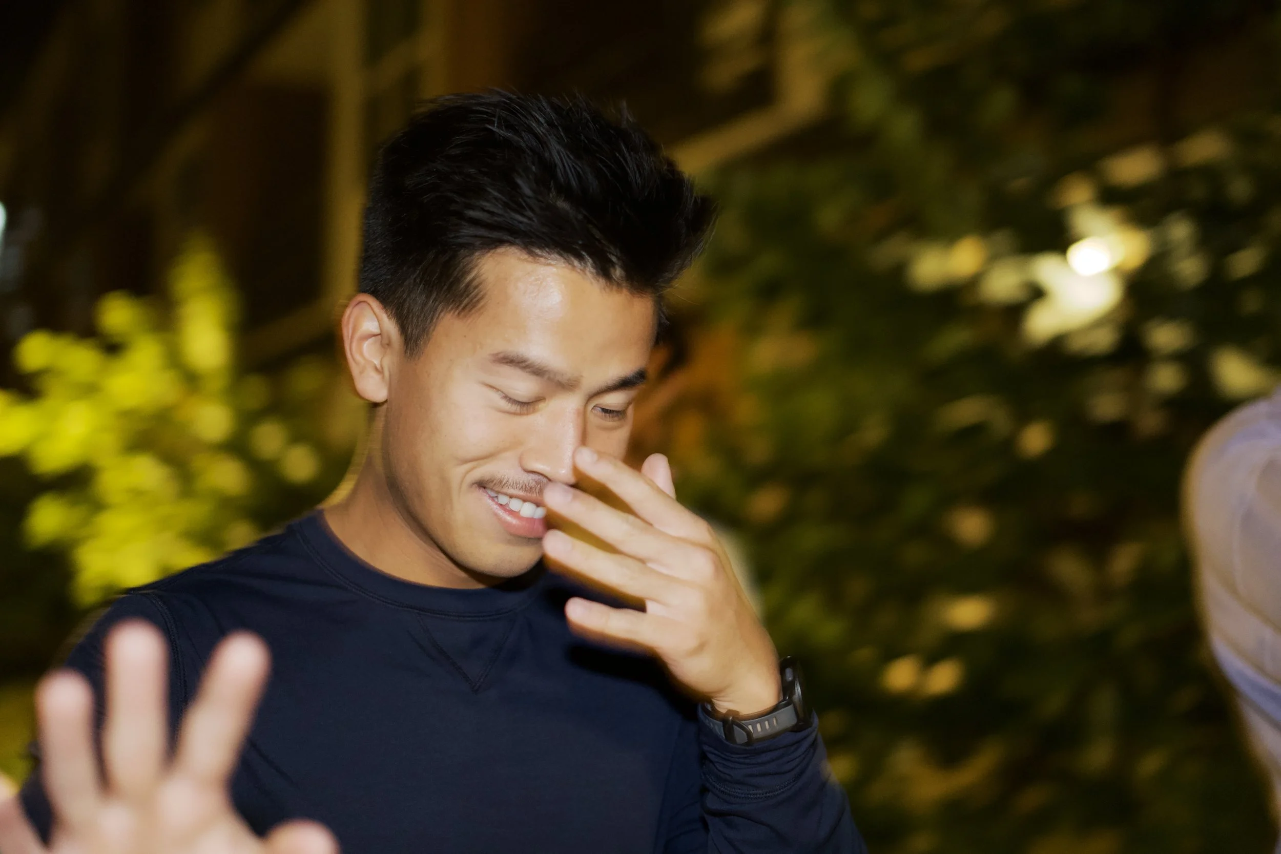 A young man with black hair and a dark shirt smiling with closed eyes, touching his face with his hand, against a background of blurred yellow-green lights.