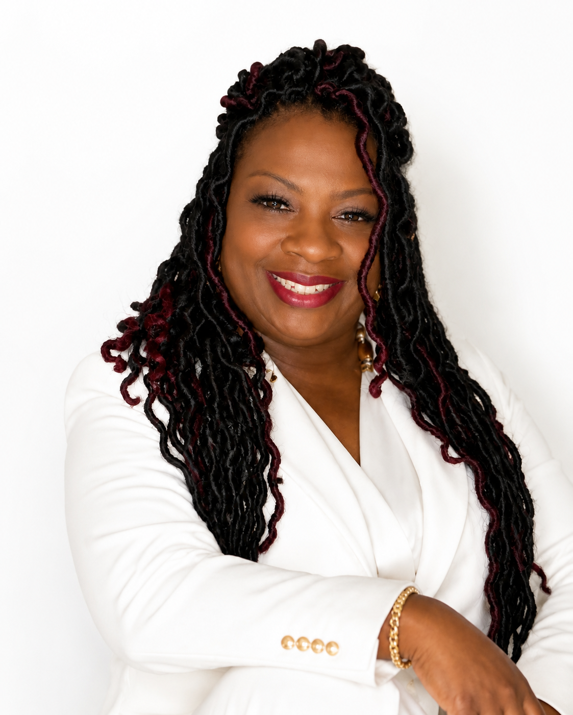 A smiling Black woman with long twisted hair in shades of black and dark red, wearing a white blazer, gold jewelry, and a red lipstick, posed against a plain white background.