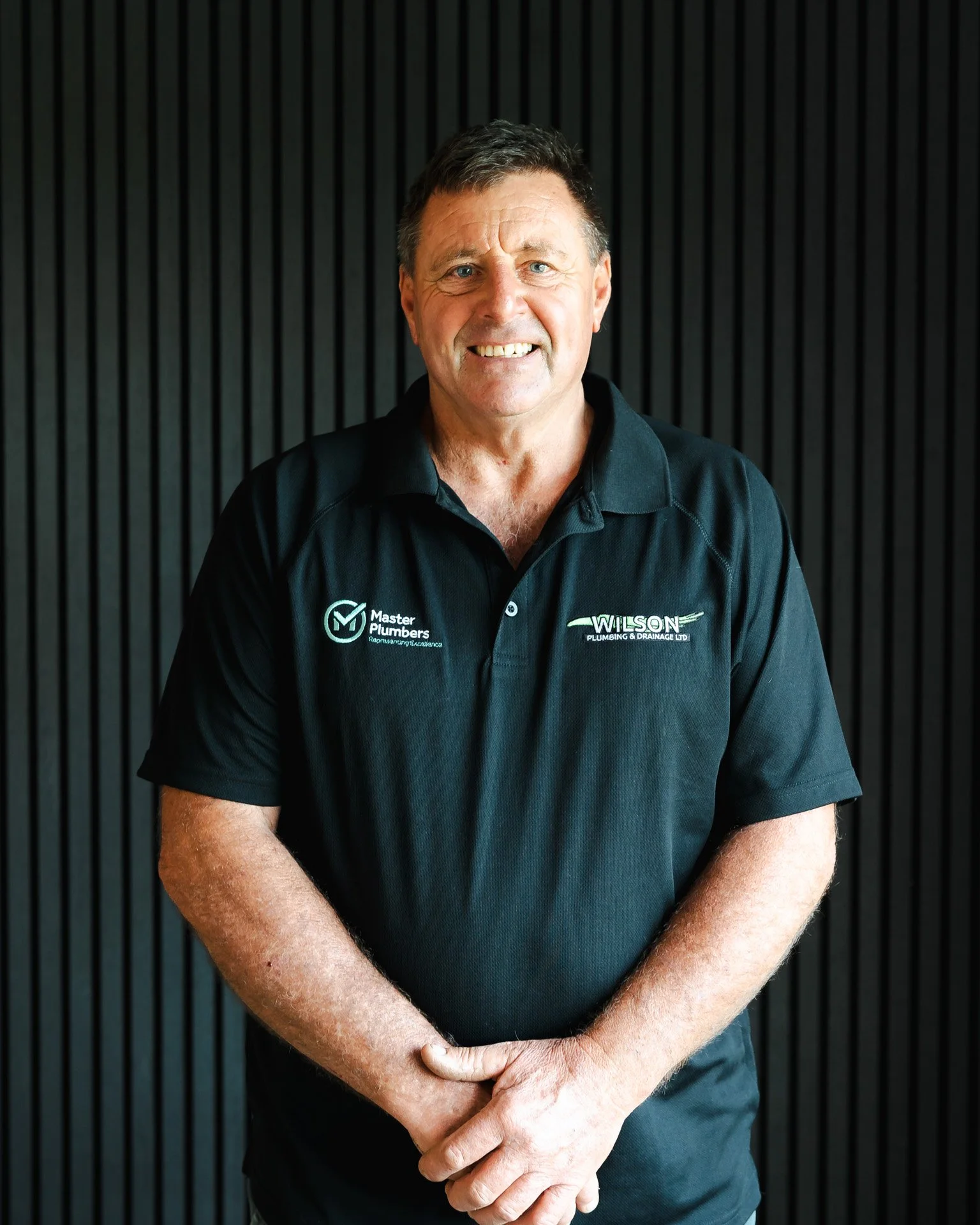 A man with short dark hair and blue eyes smiling, wearing a black polo shirt with logos and text, standing in front of a black vertical slat wall.