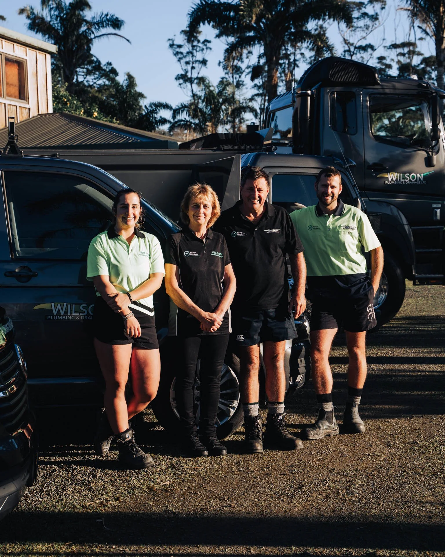 A group of four people standing outdoors in front of a black vehicle with a logo for Wilson Plumbing & Drainage. They are dressed in work uniforms and appear to be smiling for the photo. The background features trees and a building.