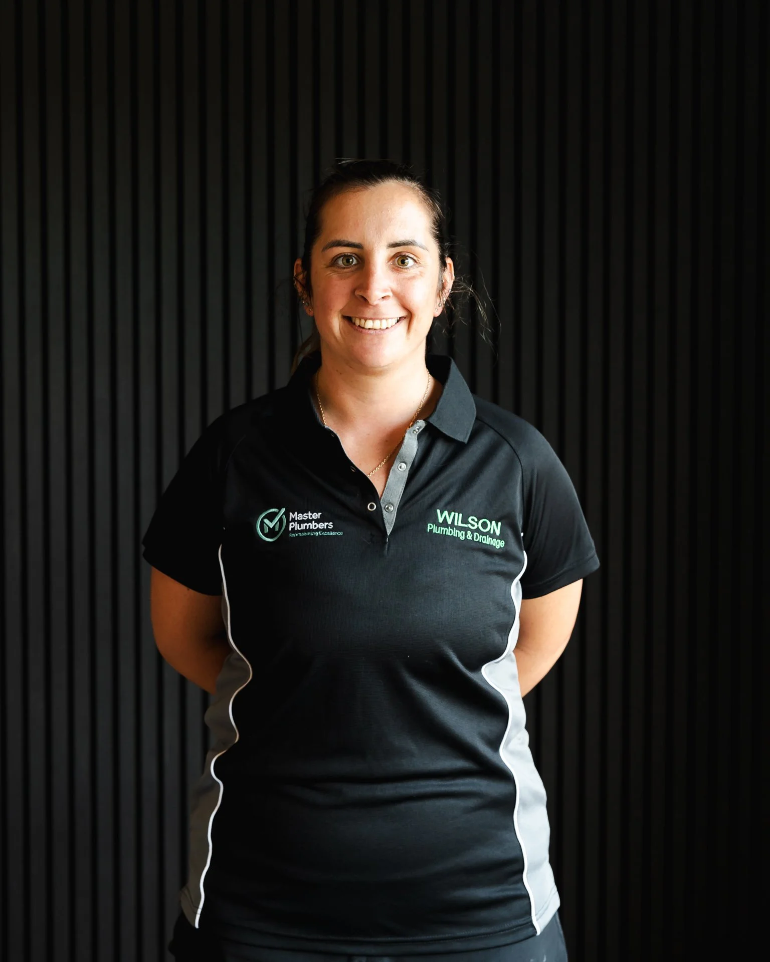 A woman with dark hair in a ponytail wearing a black and white polo shirt with company logos, standing against a black background with vertical lines.