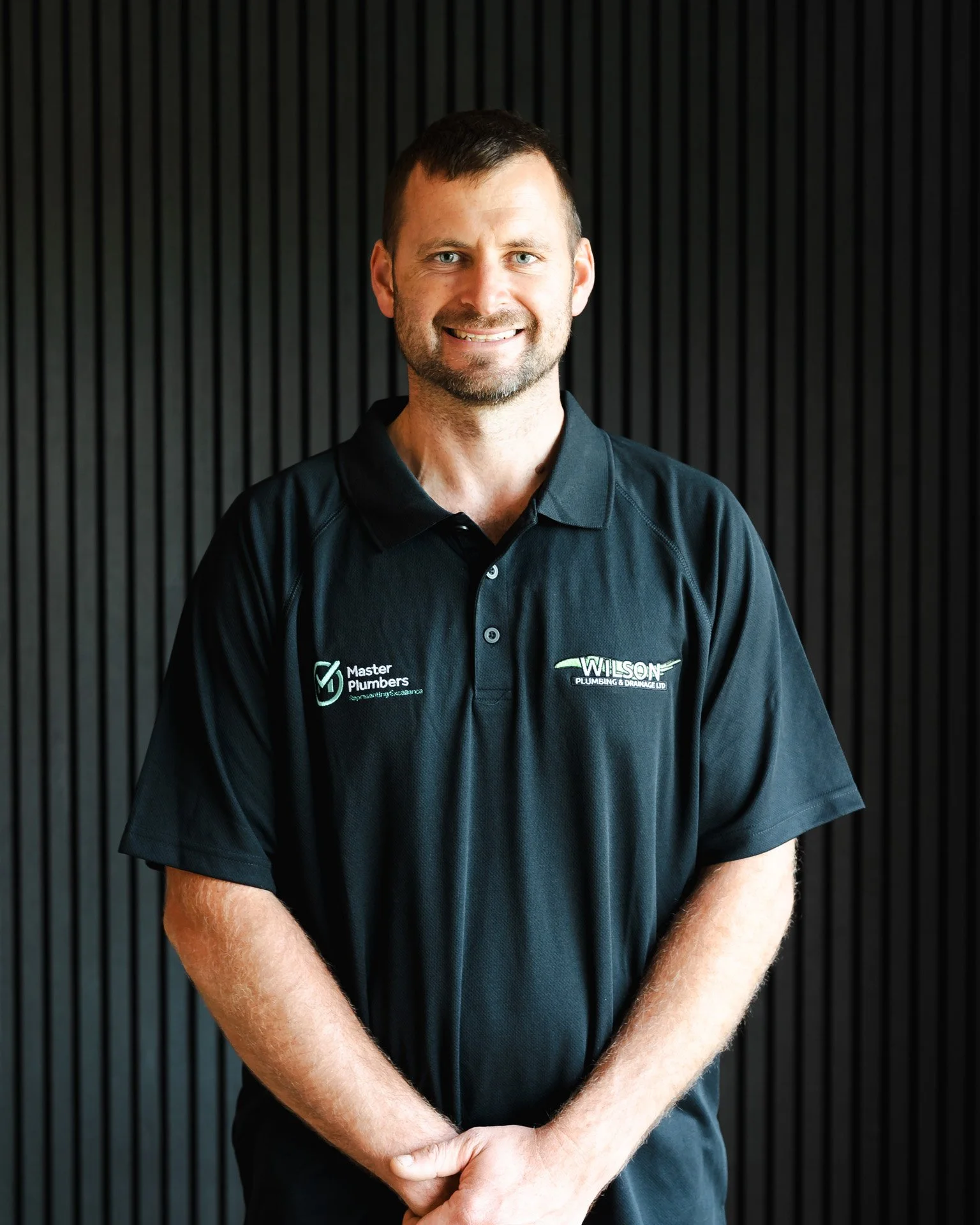 A man in a black polo shirt with company logos standing against a black slatted wall, smiling with arms crossed.