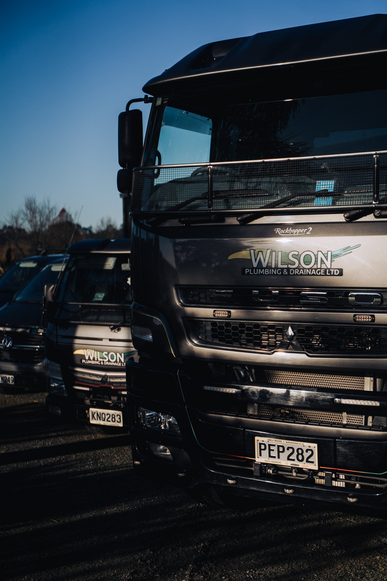 Several trucks parked in a lot with visible company logos, including Wilson Plumbing & Drainage Ltd, under a clear sky.