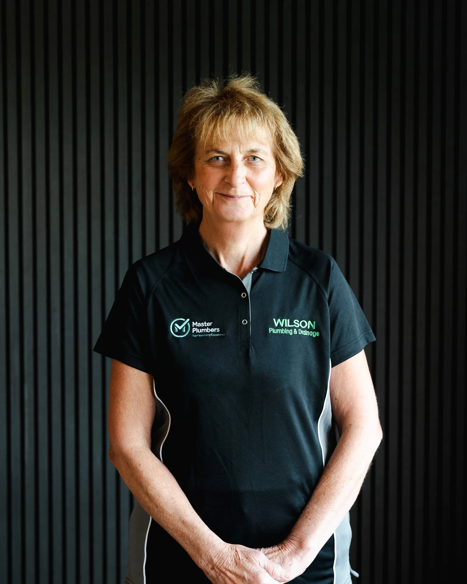 A middle-aged woman with short, light brown hair standing against a black, grooved background. She is wearing a black polo shirt with logos for "Master Plumbers" and "Wilson Plumbing & Drainage." She has a slight smile and her hands are clasped in front of her.