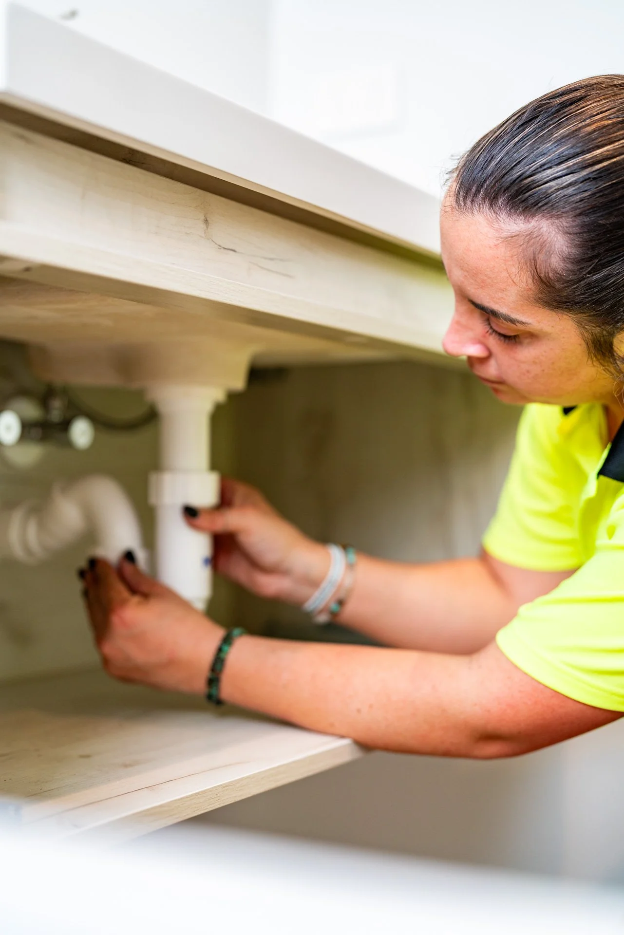 A person with long dark hair tightening plumbing pipes under a kitchen sink.
