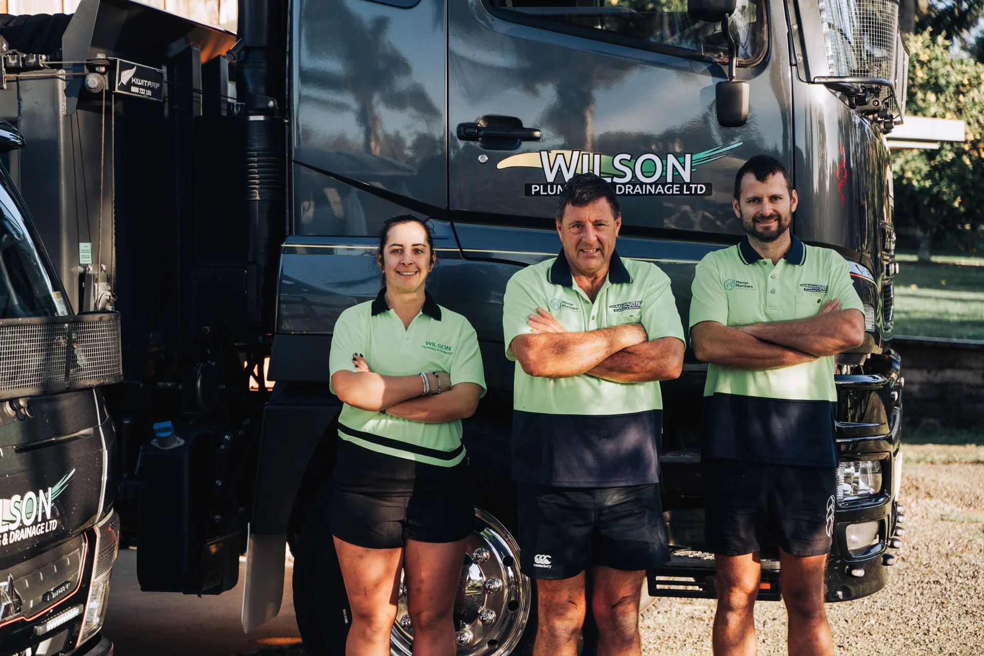 Three workers standing in front of a black Wilson Plumbing and Drainage Ltd truck, smiling with their arms crossed. The group includes two men and one woman, all wearing green Wilson uniforms. The background shows trees and a clear sky.