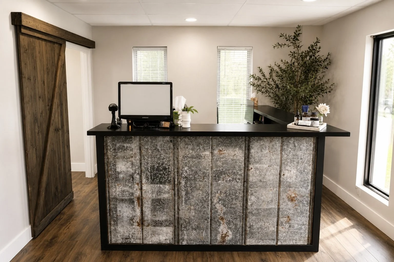 Reception desk with a rustic metal front, black countertop, computer, plant, and tissue box, in a modern office with wood flooring and a large potted plant in the background.
