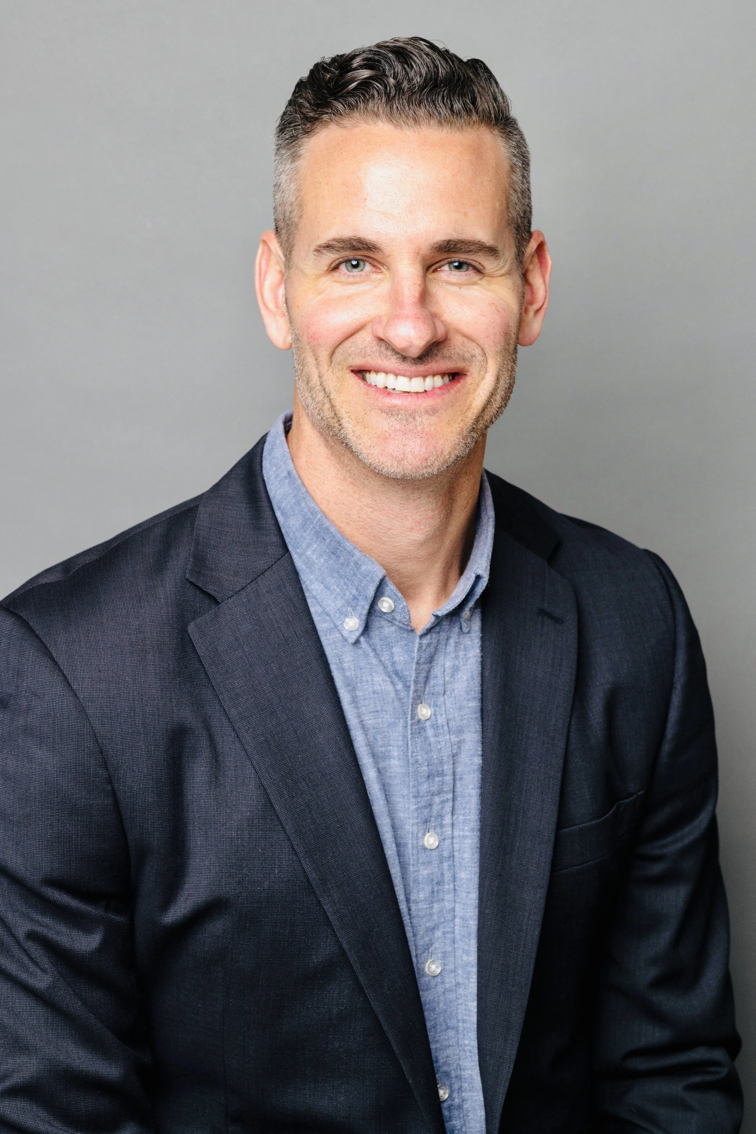 A portrait of a man with short, dark hair and blue eyes, smiling, wearing a dark blazer over a light blue button-up shirt, against a plain gray background.