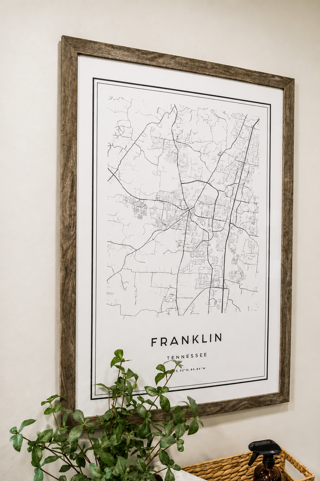 A framed map of Franklin, Tennessee, hanging on a white wall, with a green plant in front and a spray bottle in a wicker basket beneath.