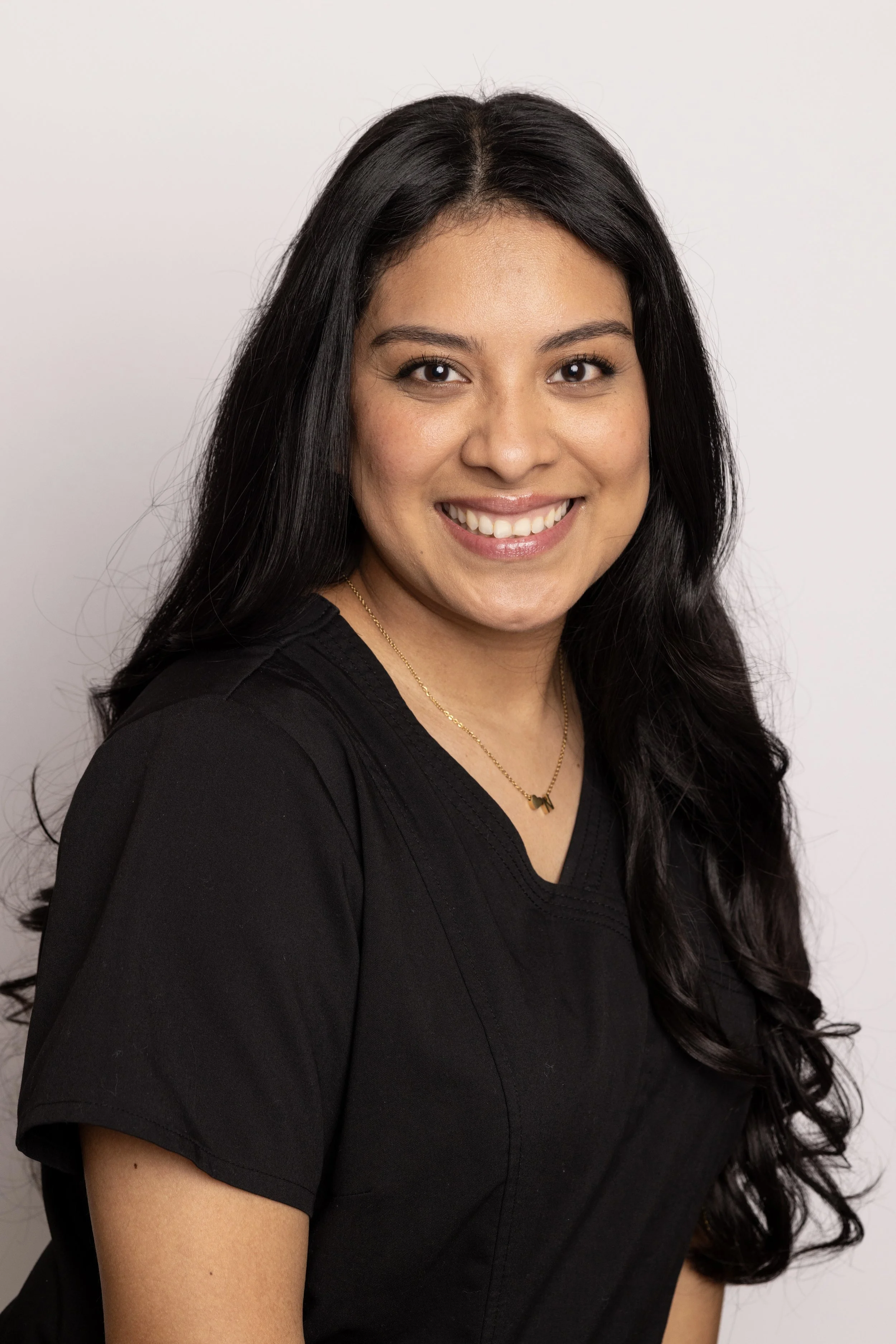 A smiling woman with long black hair, wearing a black top and a gold necklace, standing against a plain white background.