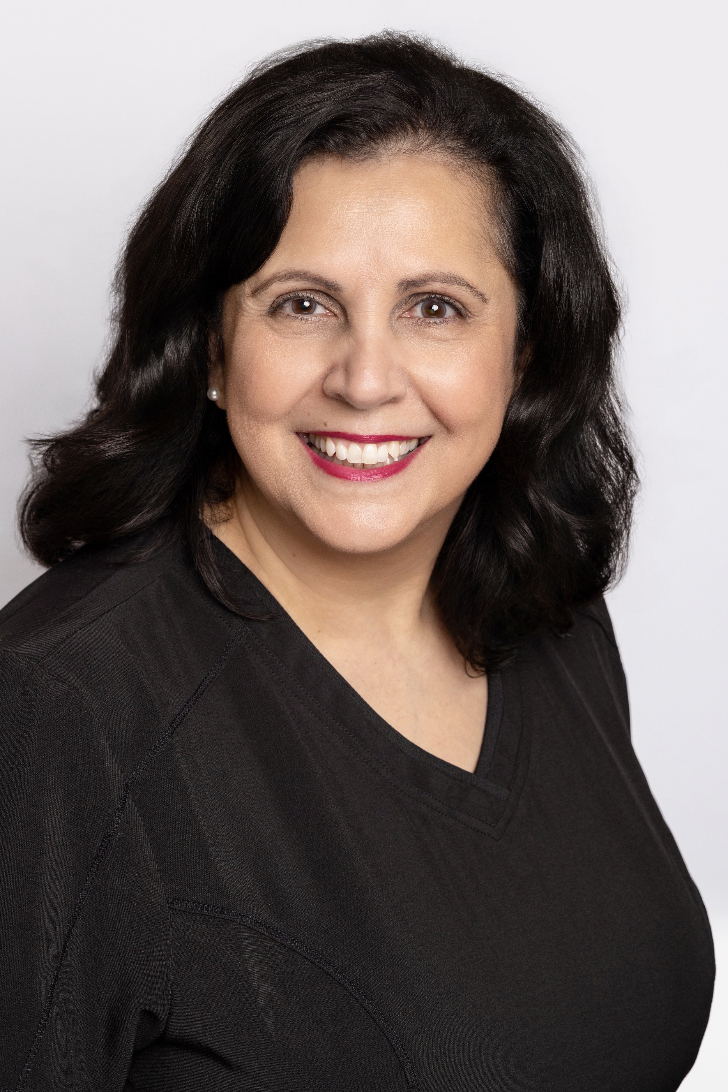 A smiling woman with dark wavy hair, wearing a black top and pearl earrings, against a light gray background.