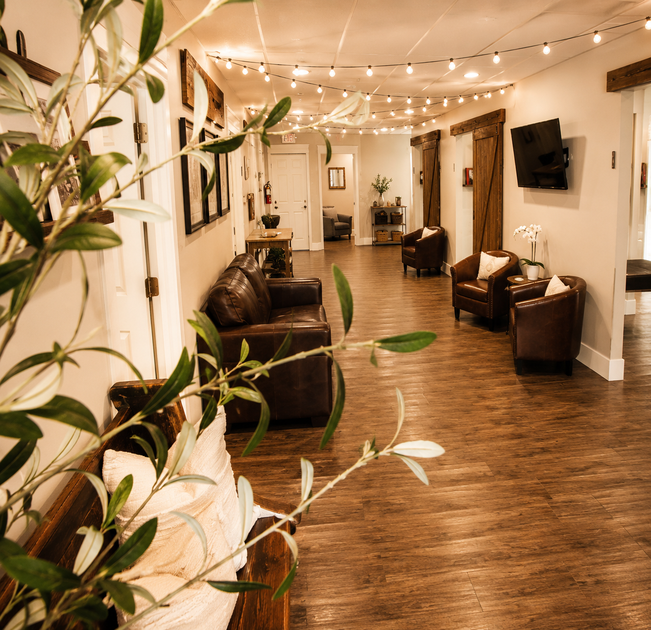A cozy waiting area with wooden flooring, leather chairs, wall-mounted TV, and warm string lights.