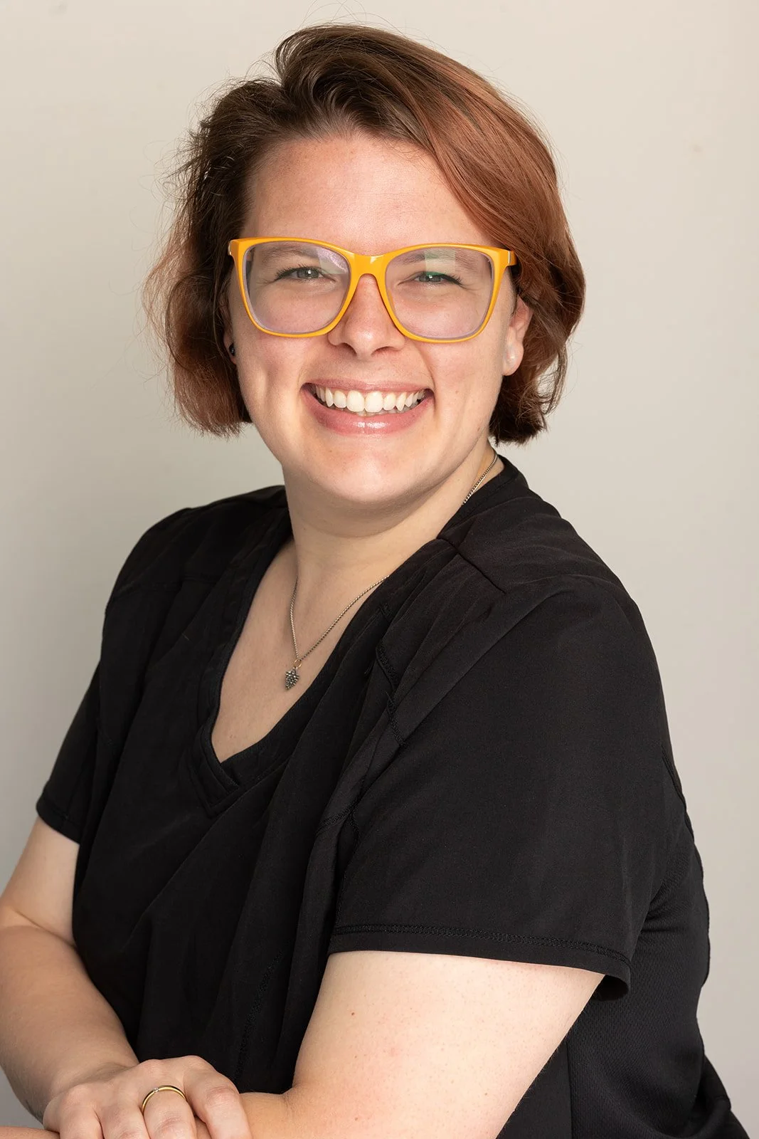 A woman with short, reddish hair and yellow glasses smiling, wearing a black shirt and a necklace, standing against a neutral background.
