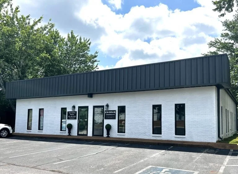 White brick building with black trim, labeled 'EXODUS' and 'Consulting' on the front, and several windows. Parking lot in front with a white car parked to the side. Trees and cloudy sky in the background.