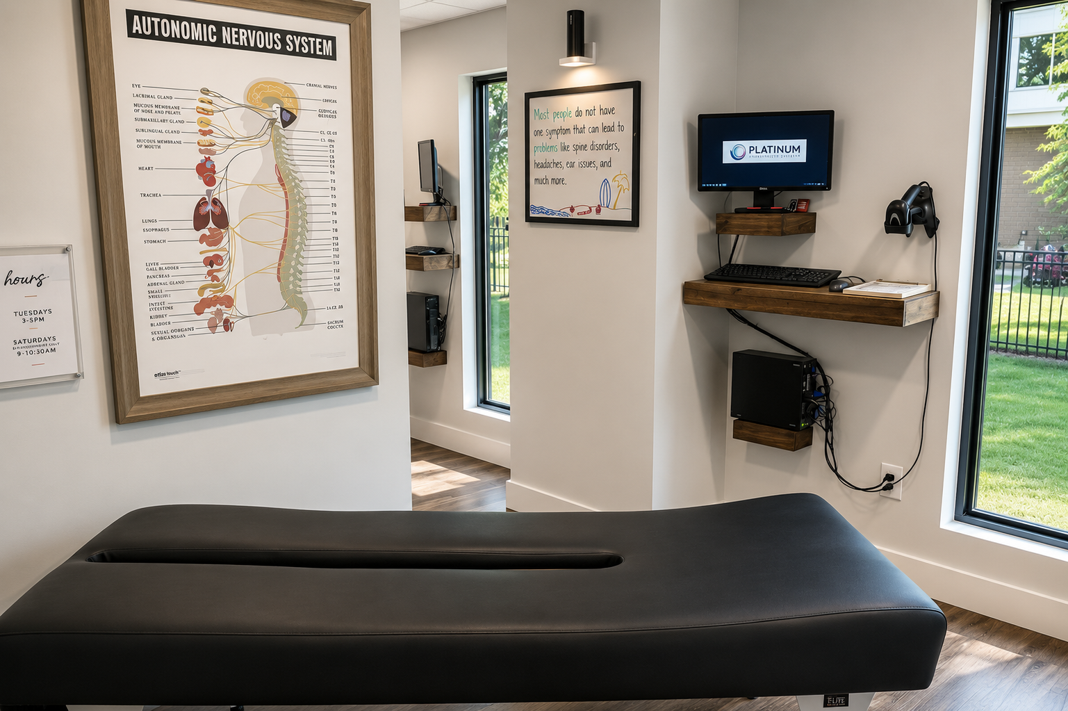 Interior of a medical office with a chiropractic table in the foreground, a large diagram of the autonomic nervous system on the wall, and a computer station with monitors and shelves on the right side.