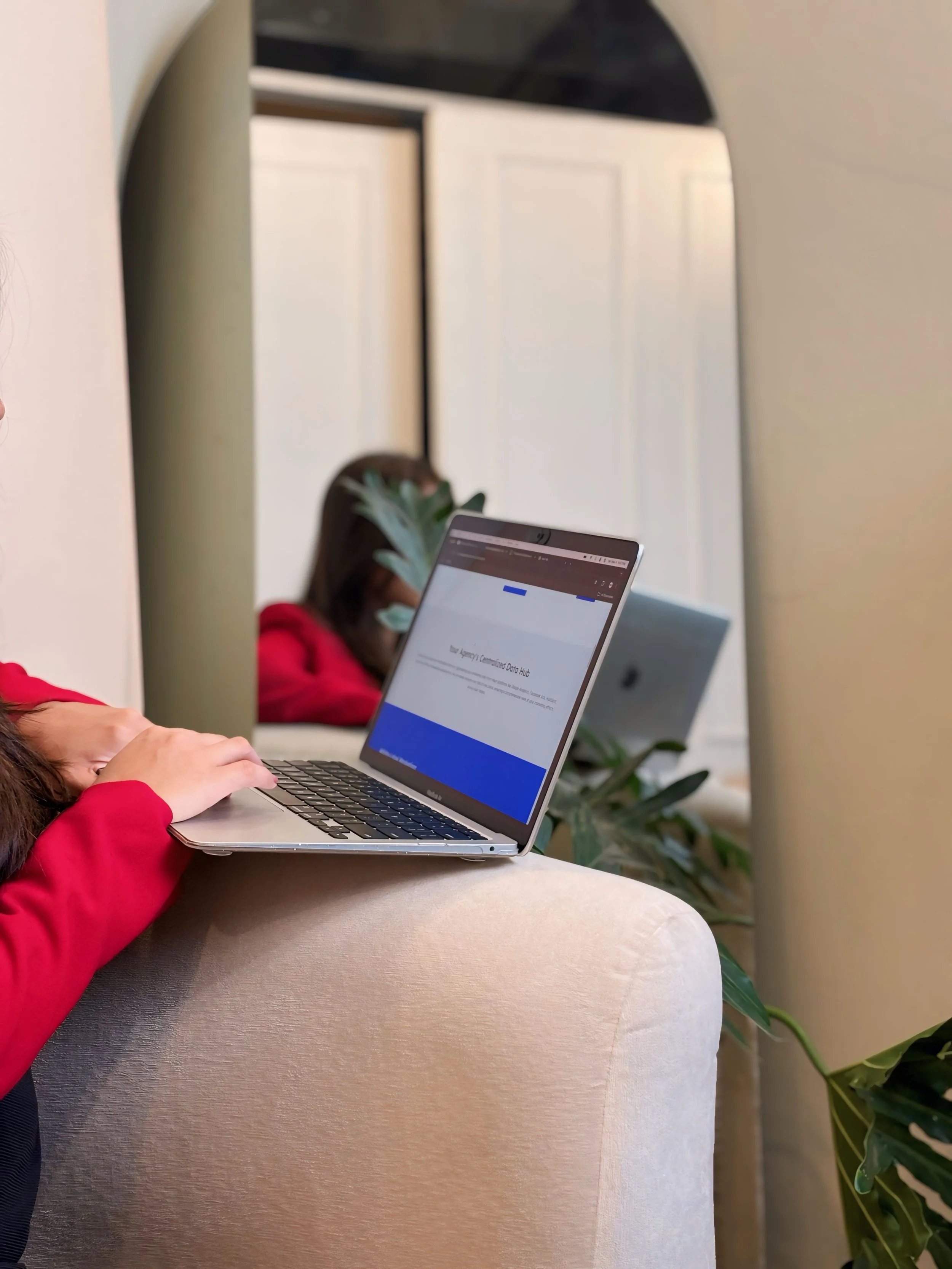 A person in a red sweater using a laptop on a beige sofa, with another person in a red sweater and plant in the background, reflected in a mirror.