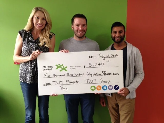 Three people standing together holding a large check, with a green and red wall in the background. The check is made out to the TWT group for $5,340, dated July 12, 2016.