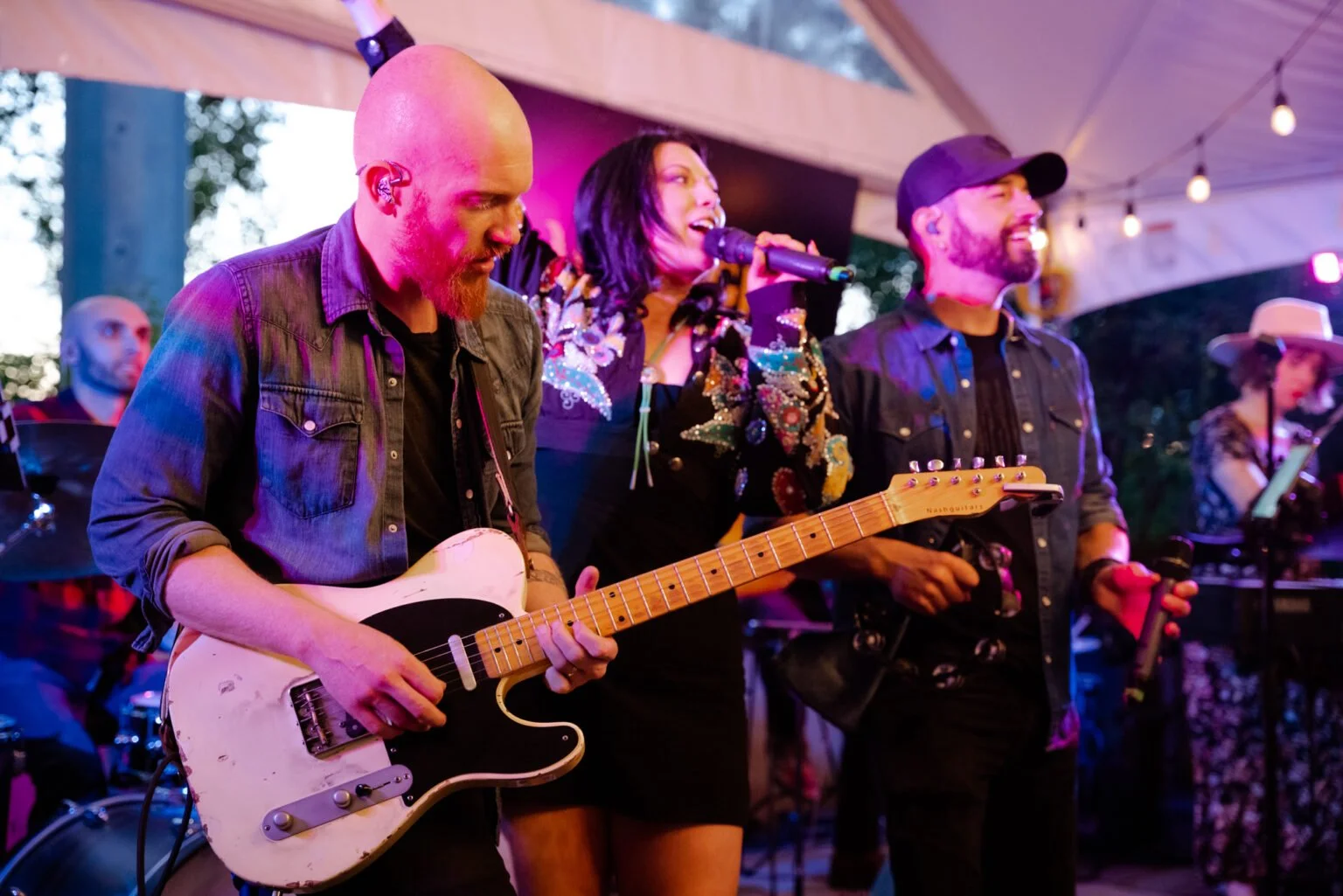 A group of musicians performing on stage at an outdoor event, with a woman singing into a microphone, a man playing electric guitar, and another man with a microphone, all under string lights.