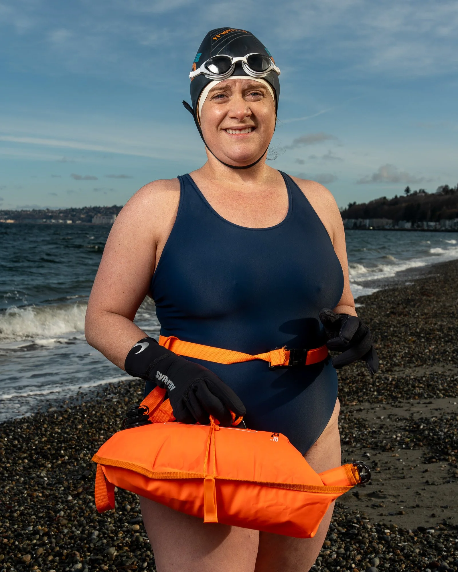 A woman standing on a rocky beach in a swimsuit, wearing a swimming cap and goggles, holding an orange life jacket, with water and a distant shoreline in the background.