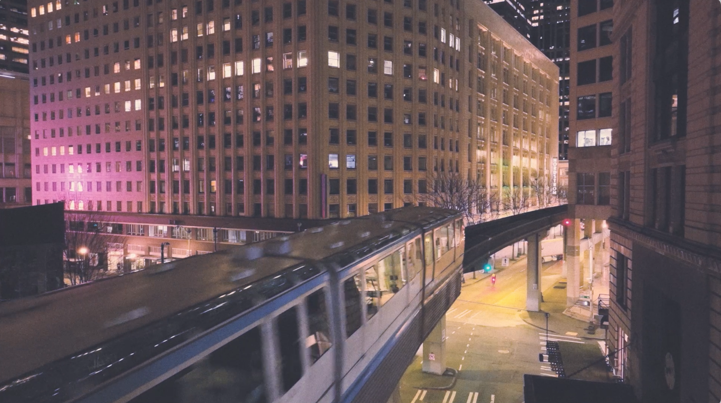 Nighttime city scene with tall buildings, a monorail train on an elevated track, and street lights.