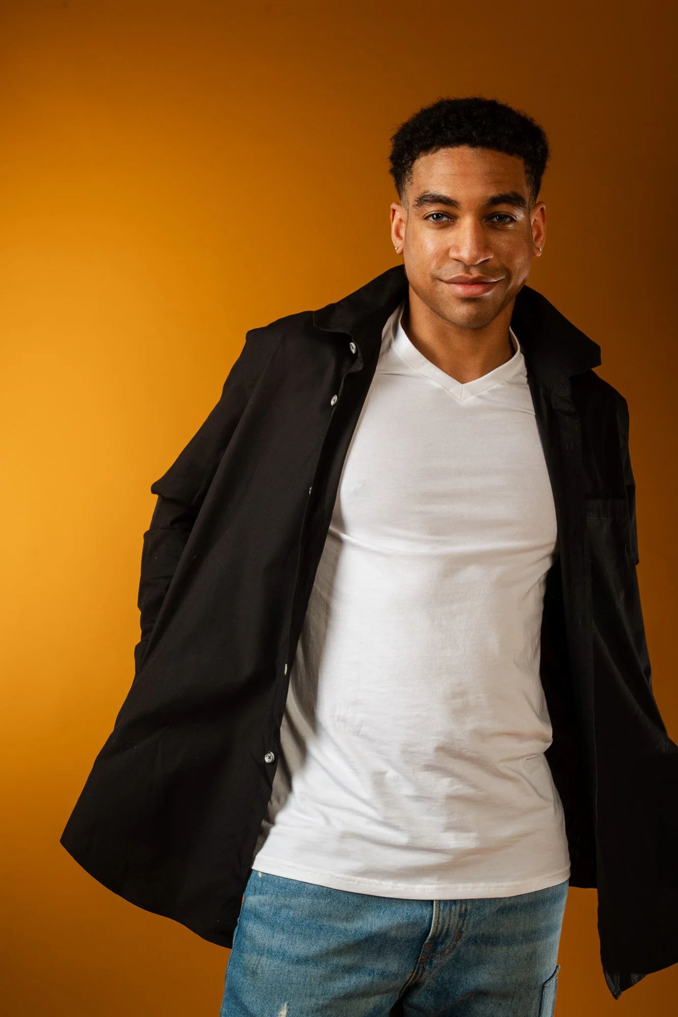 A young man with short curly dark hair and earrings smiles lightly at the camera. He wears a black jacket over a white shirt and blue jeans, standing against a warm orange background.