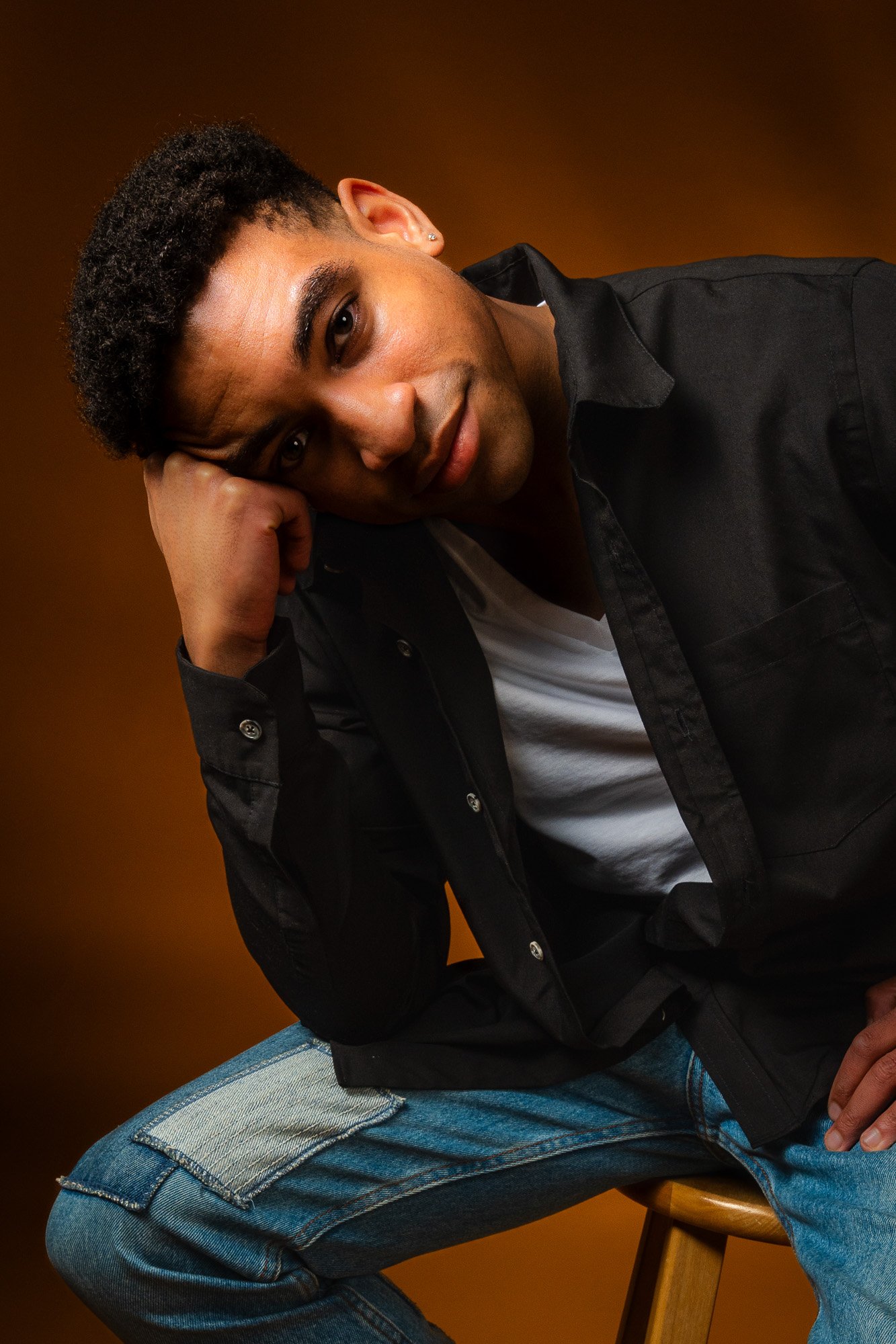 A young man with curly hair, earrings, and a black shirt sitting on a wooden stool against a brown background.