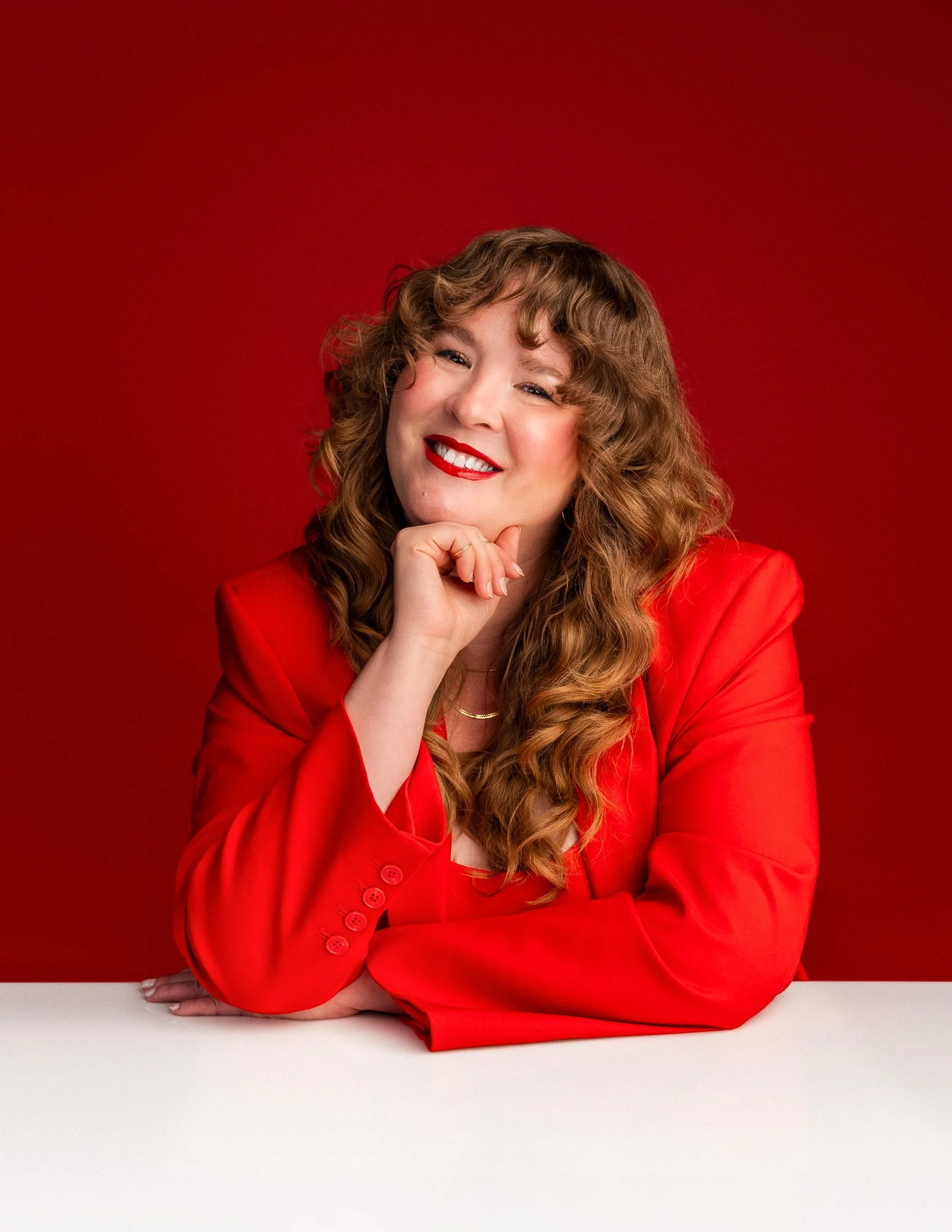 A woman with curly hair and red lipstick wearing a red blazer, smiling with her chin resting on her hand, against a red background.
