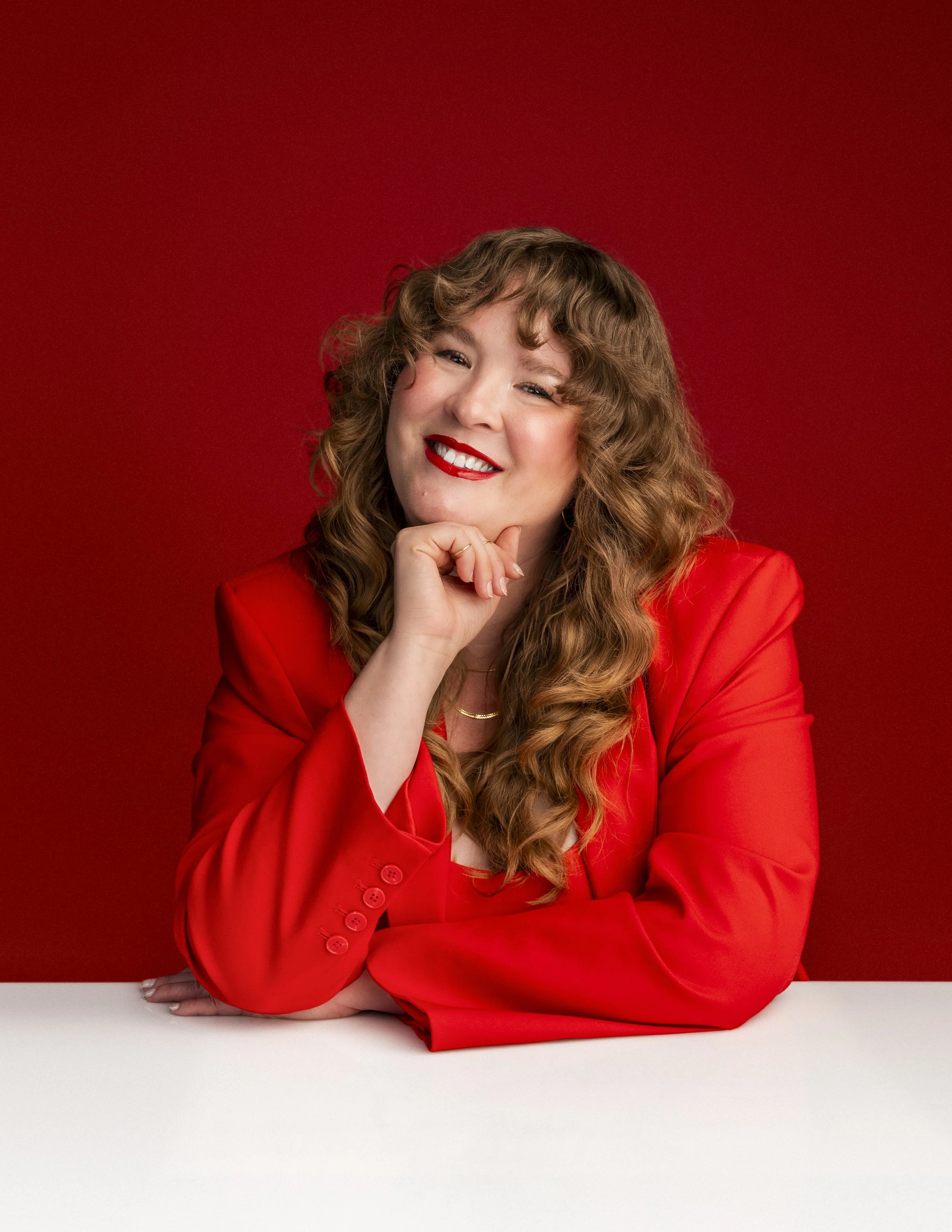 A woman with curly hair wearing a red blazer, sitting at a white table with a red background, smiling and resting her chin on her hand.