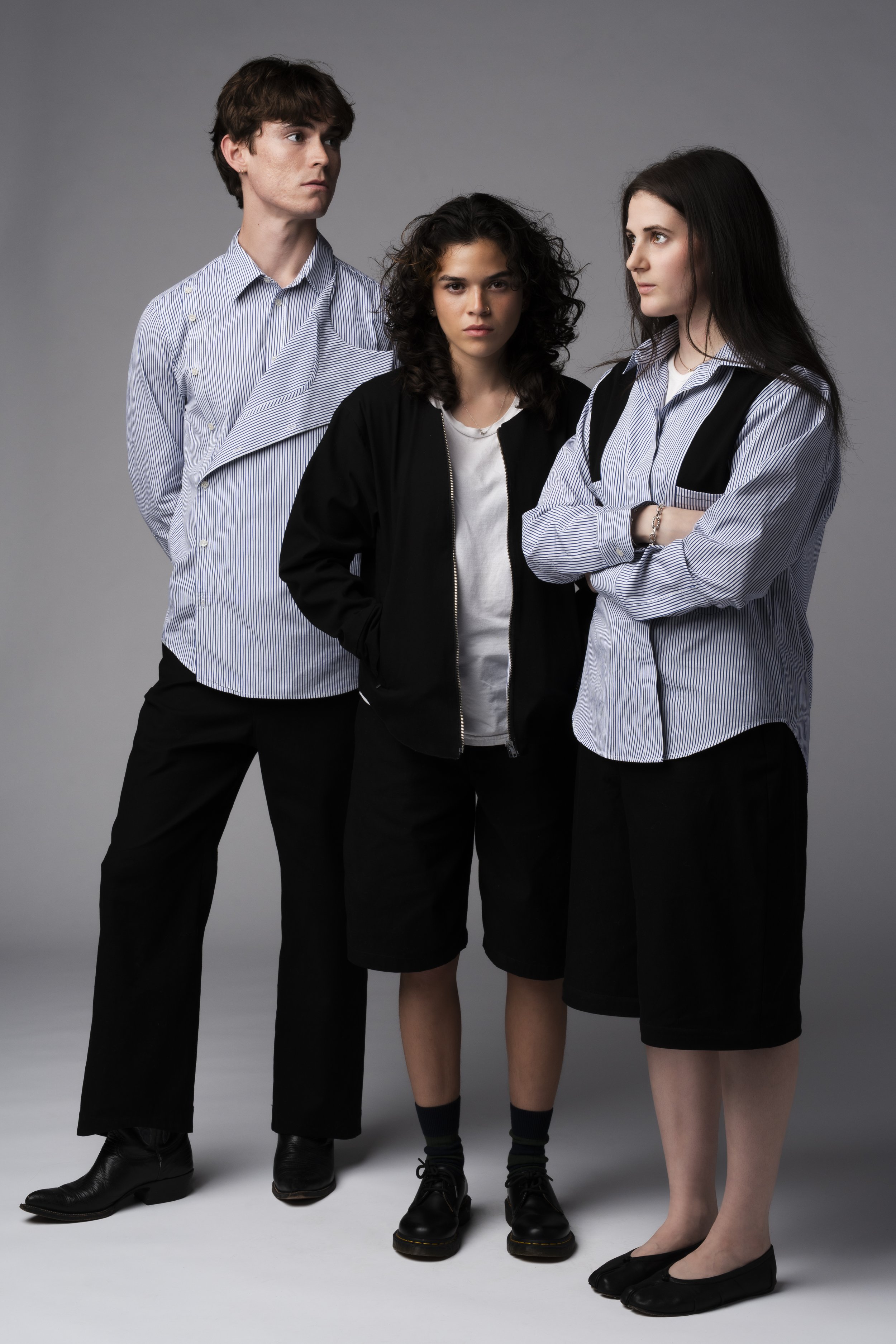 Three young women standing close together against a neutral gray background, dressed in casual clothing.