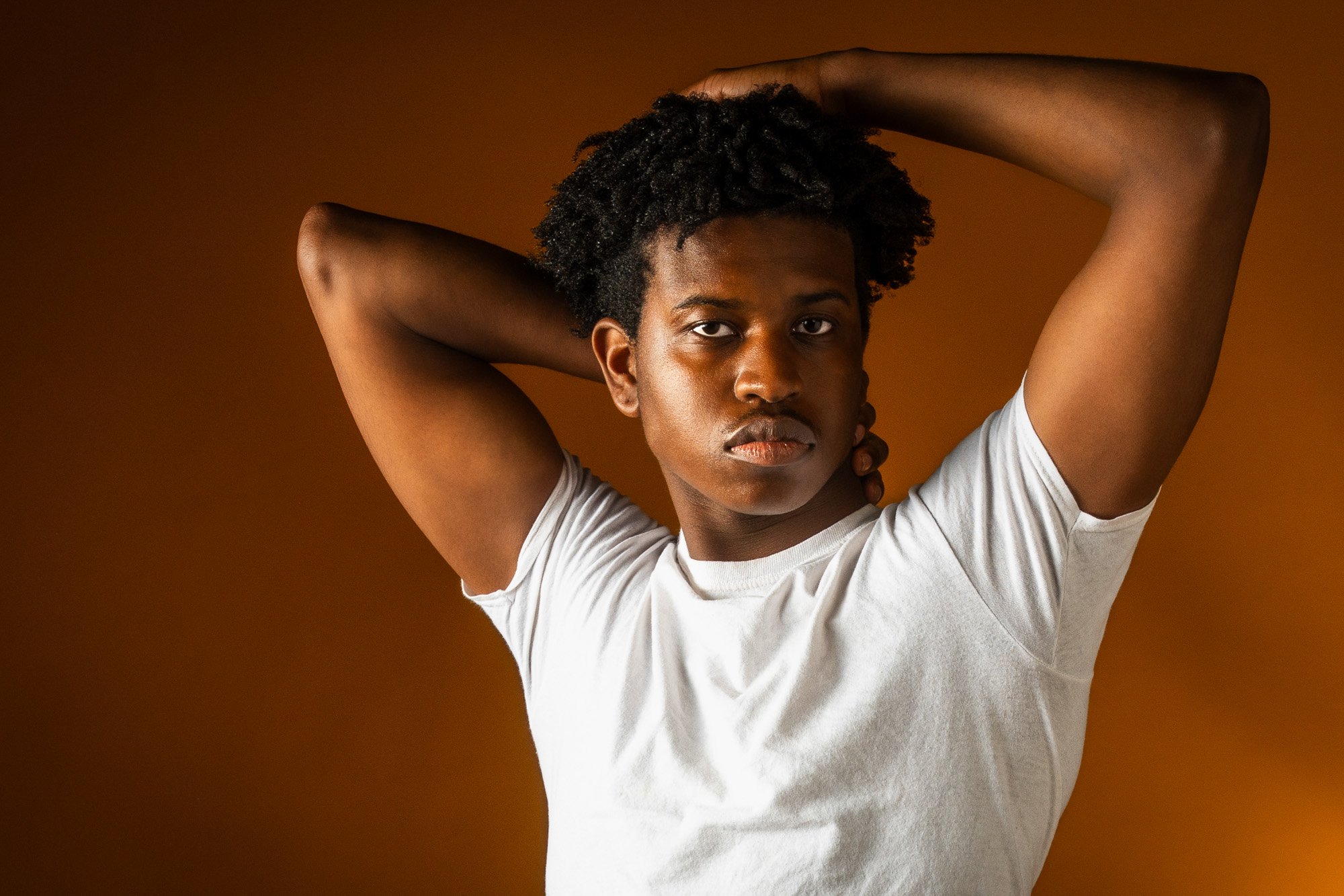 Young person with dark, curly hair and tan skin, wearing a white t-shirt, with arms raised and hands behind their head, standing against a warm brown background.