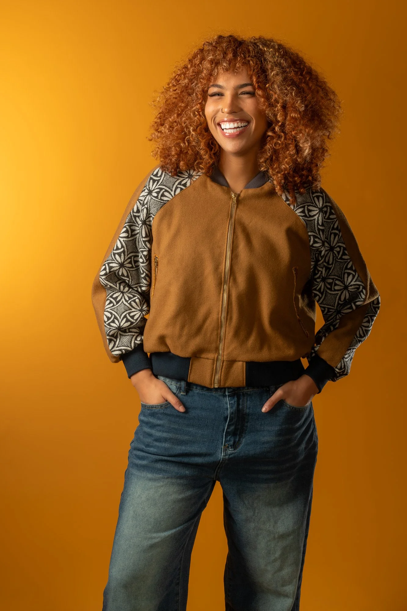 A woman with curly reddish-brown hair, smiling, wearing a brown bomber jacket with patterned sleeves, jeans, standing in front of a yellow background.