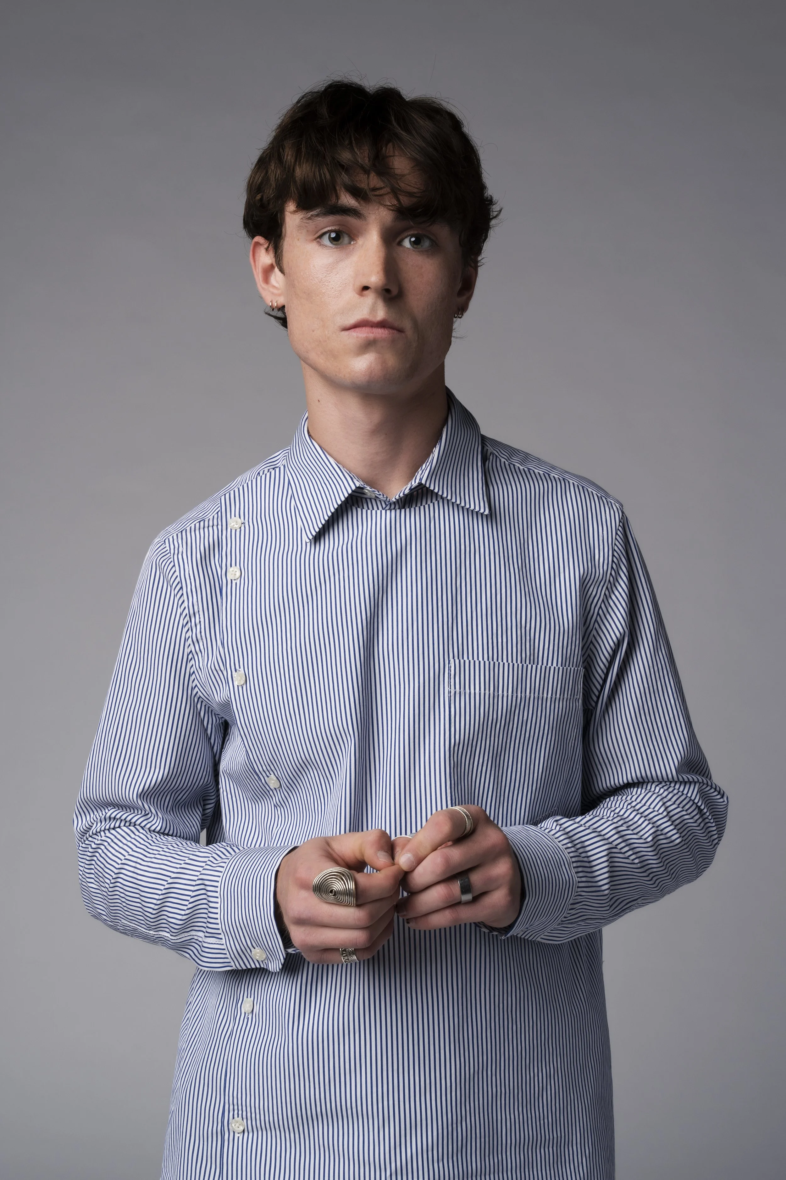 A young man with short brown hair, wearing a blue and white striped shirt and multiple silver rings, standing against a plain gray background.