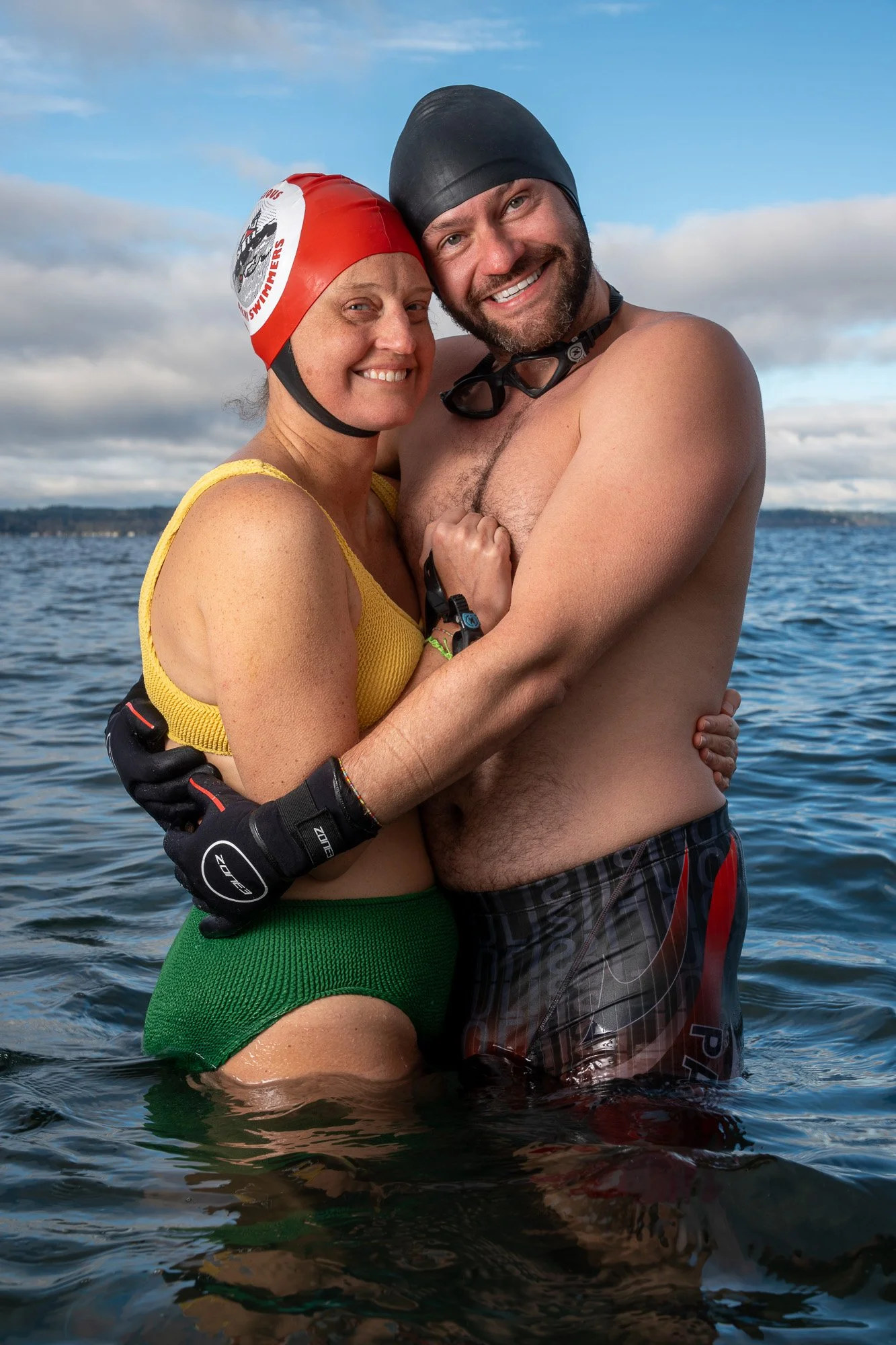 A smiling man and woman are embracing in water, wearing swimming gear and goggles, with a cloudy sky and distant shoreline in the background.