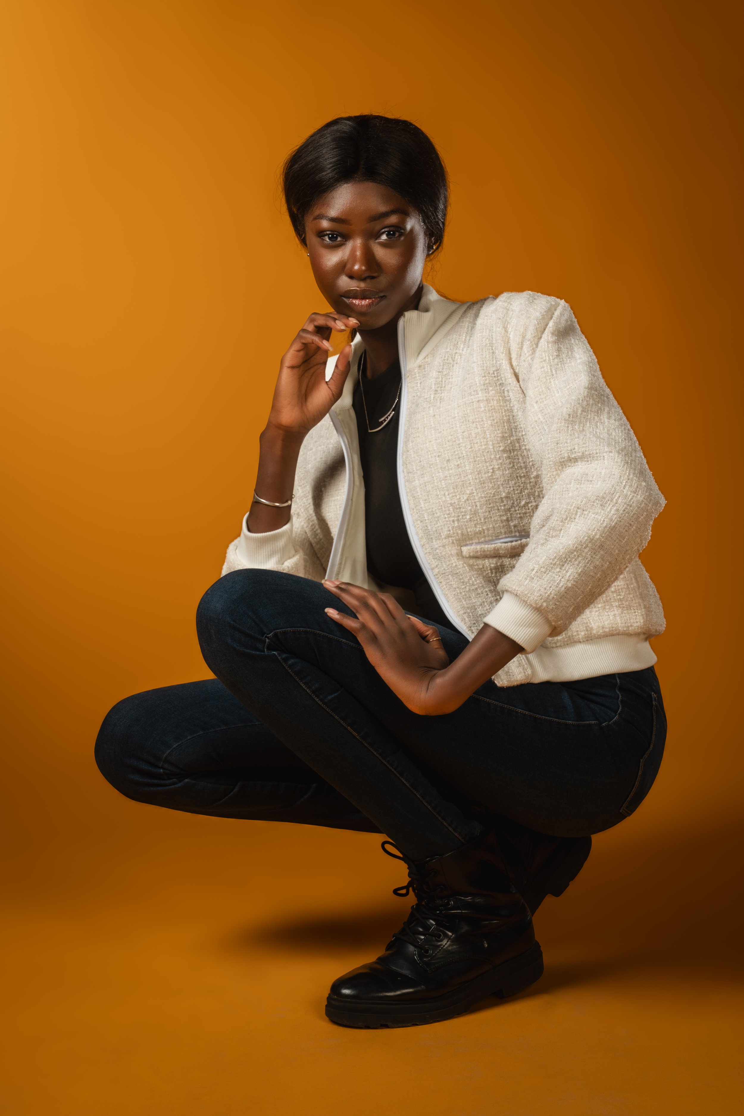 A young woman with dark skin, short black hair, wearing a beige jacket, black shirt, dark jeans, and black boots, is posed against an orange background. She is squatting with one knee up and her hand resting on her chin.