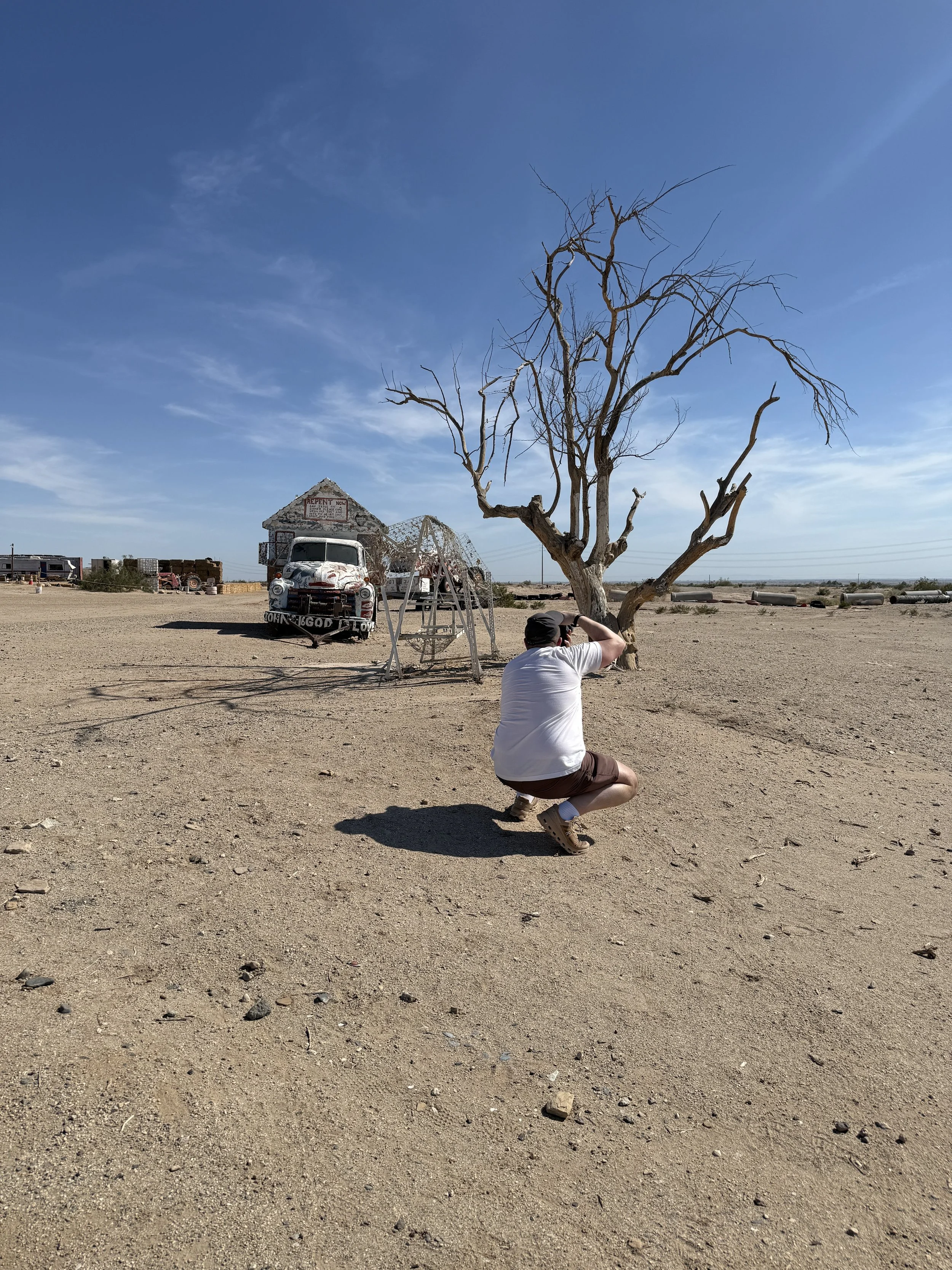 A man kneeling in a desert landscape, taking a photo of an old, leafless tree with twisted branches. There is a rusty, vintage car and a dilapidated building in the background, under a clear blue sky.