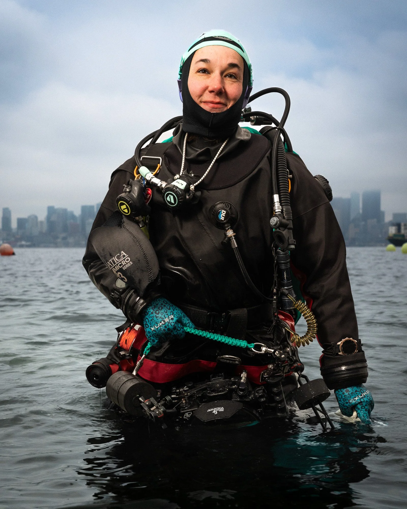A female diver standing in water, wearing a black wetsuit, diving gear, and a helmet, with a city skyline in the background.