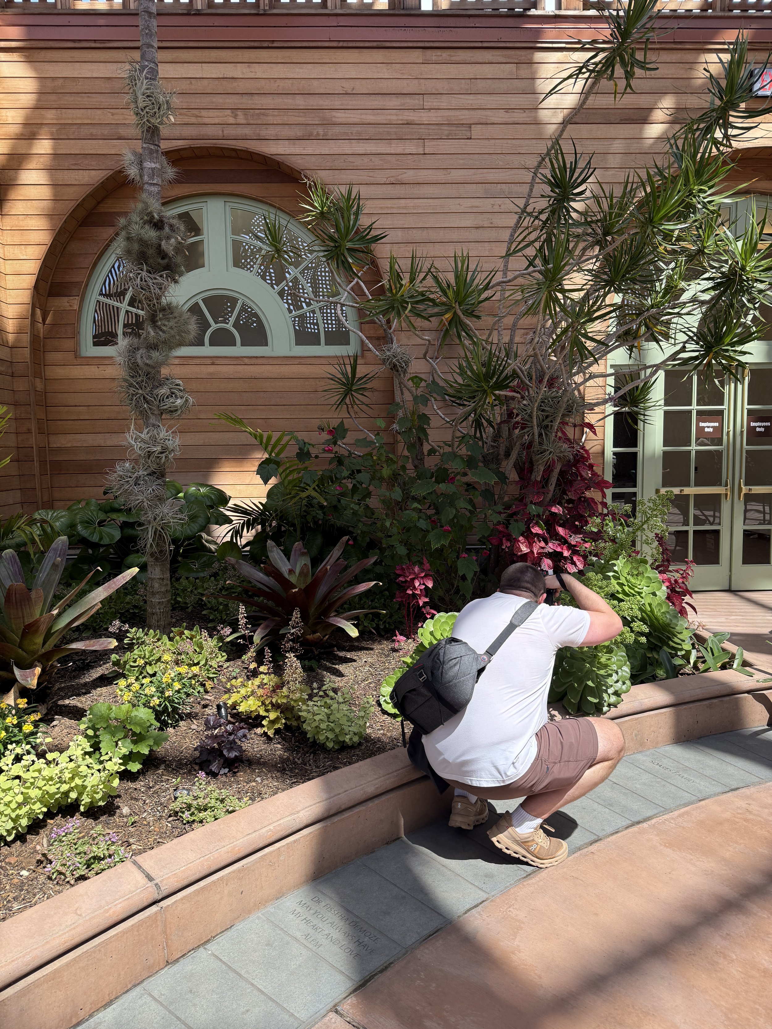 A man crouching and taking a photo of an engraved memorial plaque at a garden with lush green plants and trees, a stone walkway, and a wooden fence in the background.